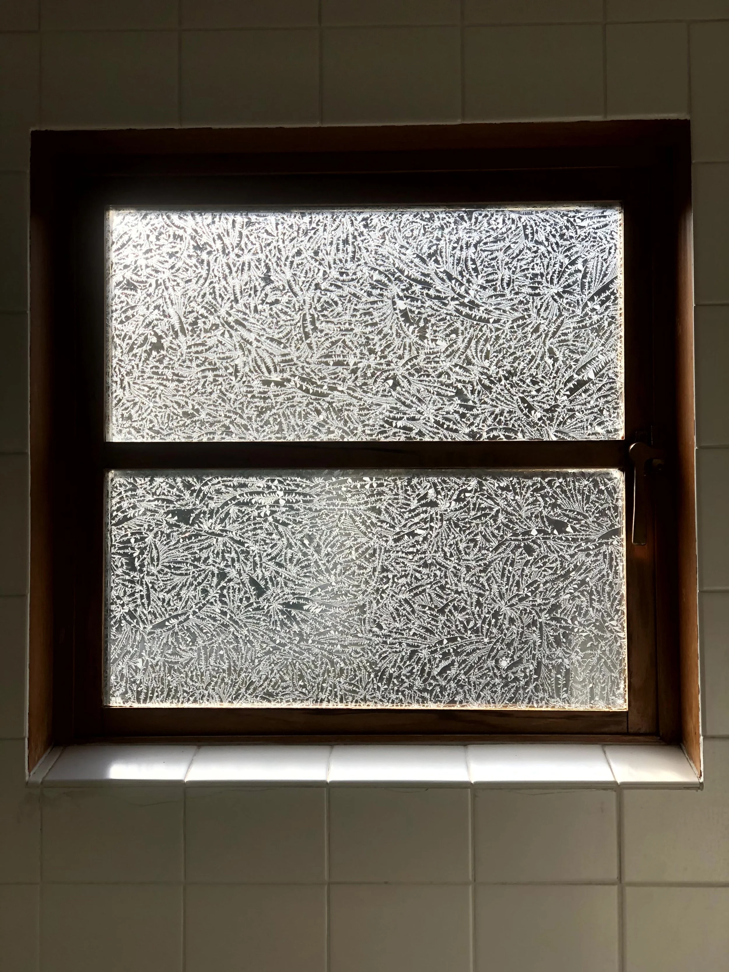 Frosted window with ice crystals forming intricate patterns, surrounded by white tiled wall.