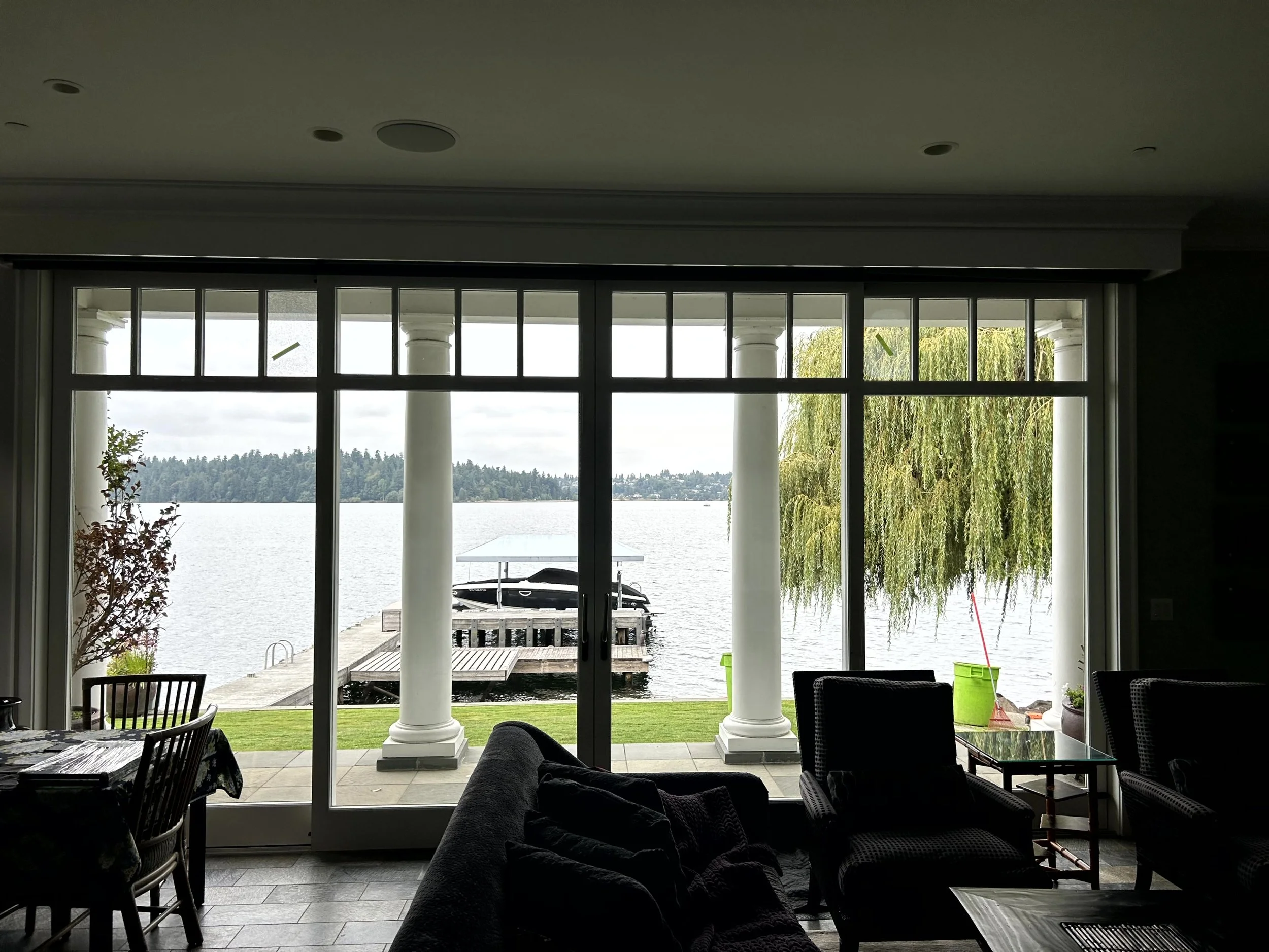 Indoor view of a house with large glass doors opening to a lakeside deck with boats, columns, lush trees, and overcast sky.