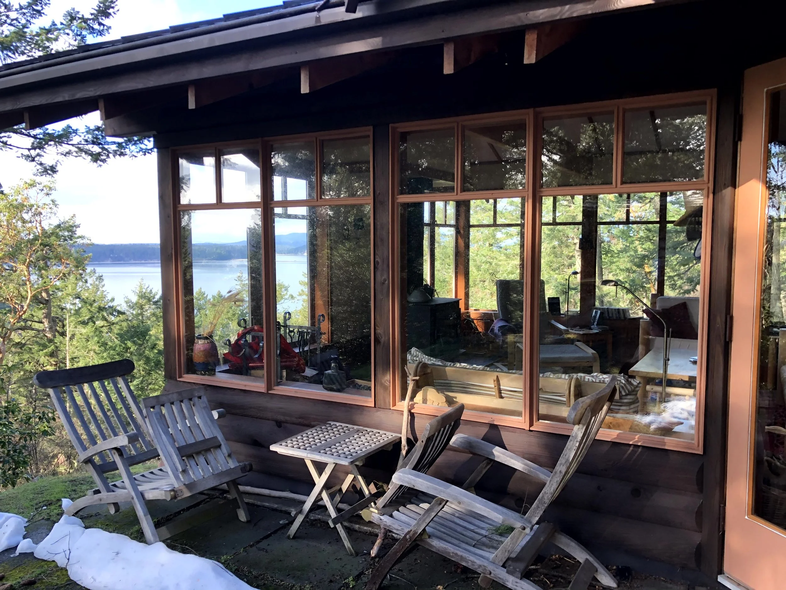 View of a rustic house with large windows overlooking a lake, surrounded by trees. Wooden outdoor furniture is arranged outside on a small patio.