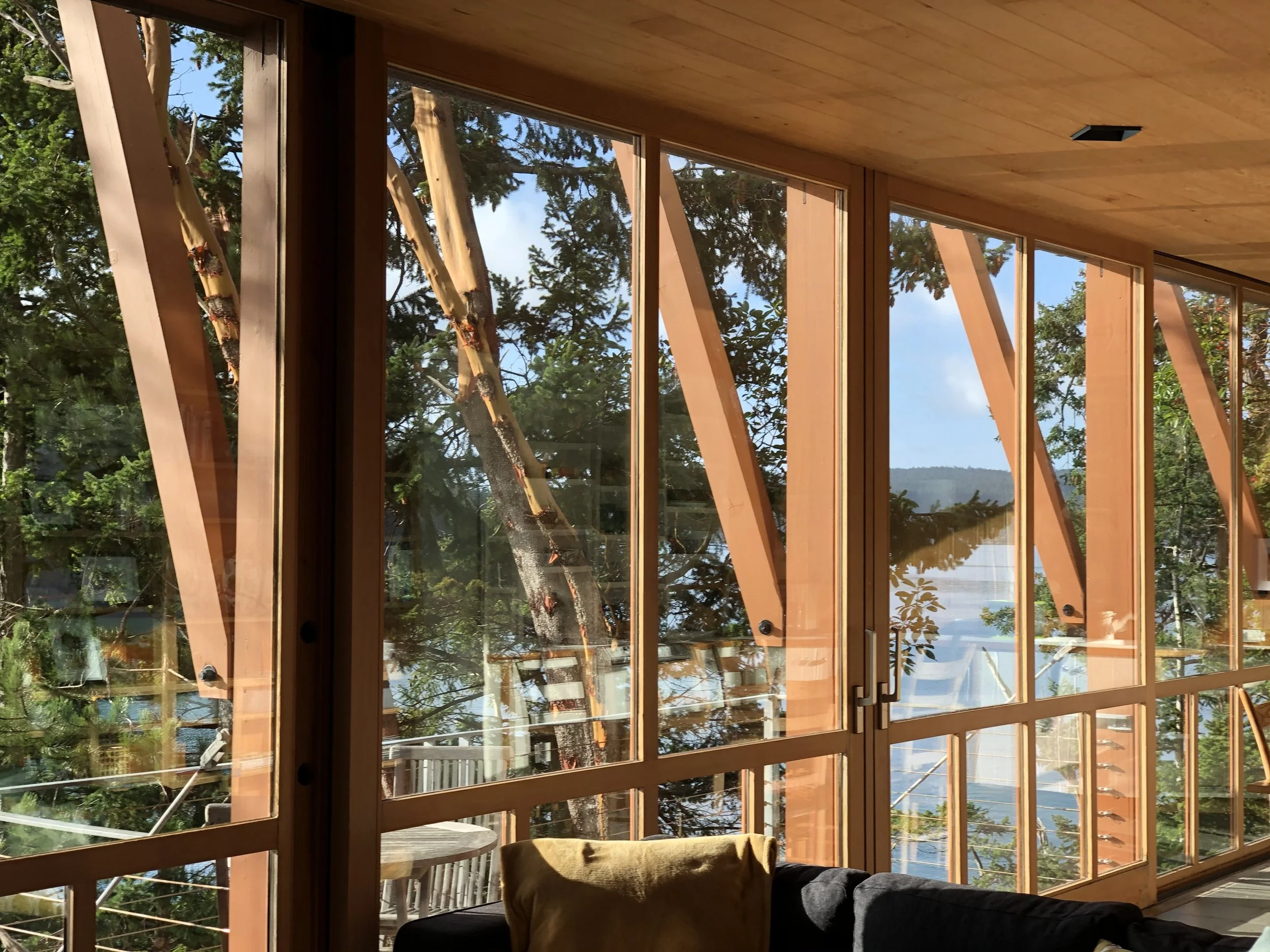Interior view of a room with large glass windows showing trees and water outside, with wooden framing and ceiling, and a sofa with pillows.