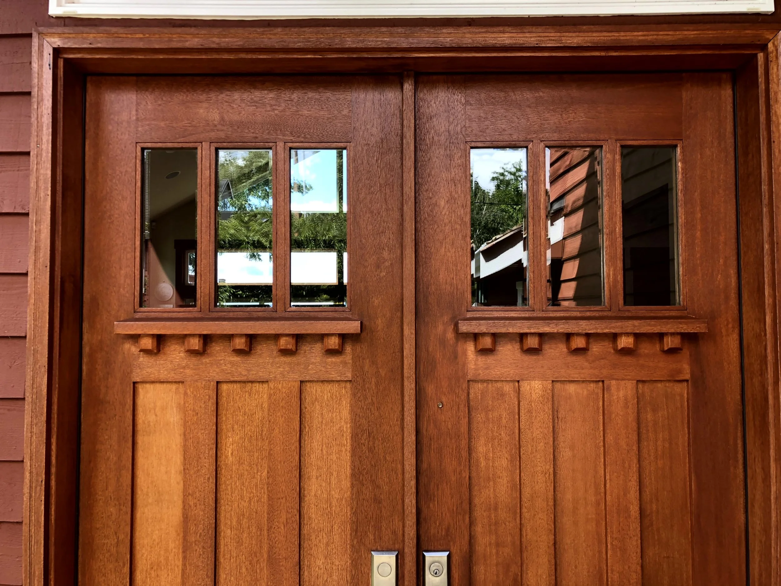 A double wooden front door with rectangular glass panes at the top, reflecting trees and the sky, set in a redwood house exterior with vertical siding.