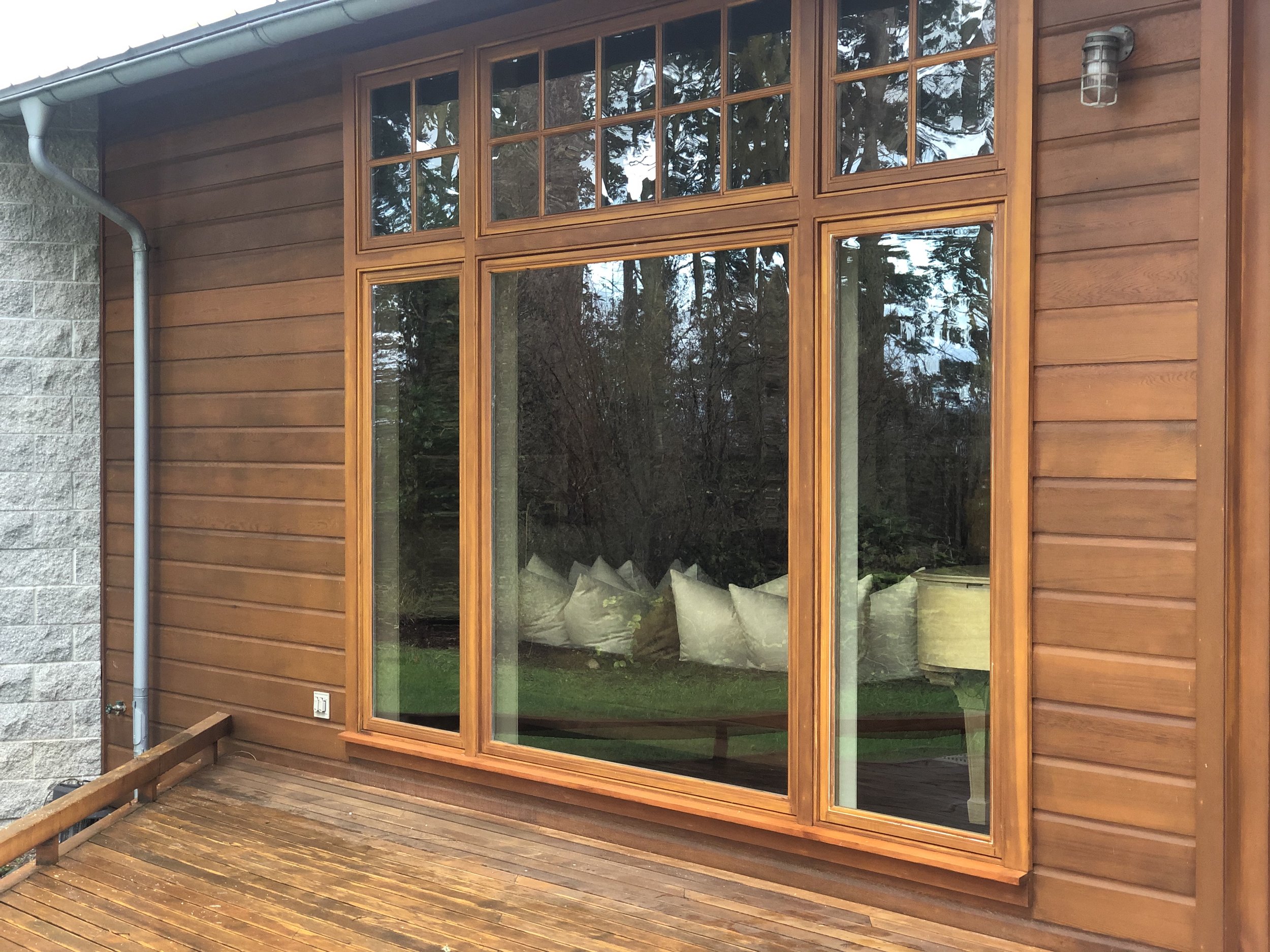 Large wooden-framed glass sliding door leading to a deck with wood flooring. The door has multiple panes and reflects outdoor trees. To the left, there is a white brick wall and a grey downspout.