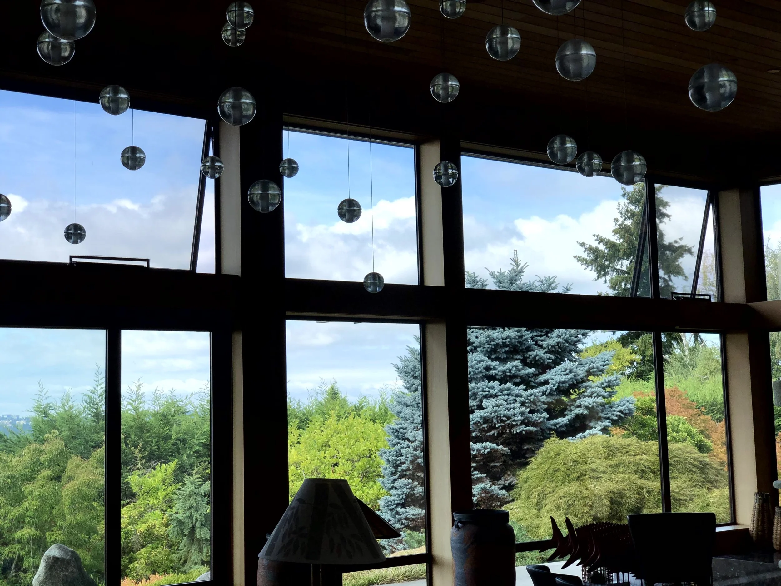 Interior view of a room with large windows overlooking lush green trees and a cloudy sky, decorated with hanging glass orbs and a table lamp.