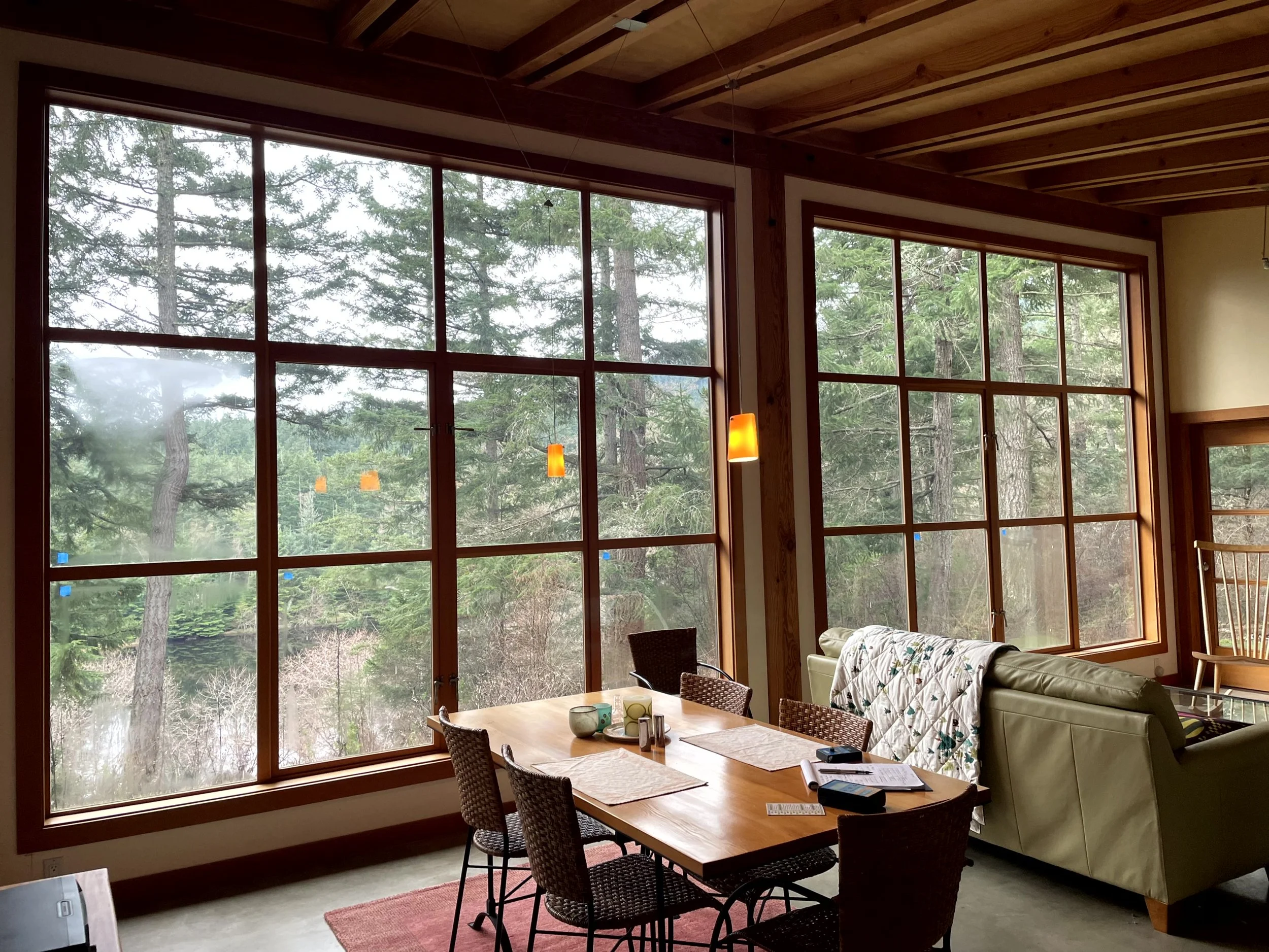 Interior view of a living/dining room with large windows showing a wooded outdoor landscape, a rectangular wooden table with chairs, a beige sofa with a quilt on the back, a lamp, and a wooden rocking chair.