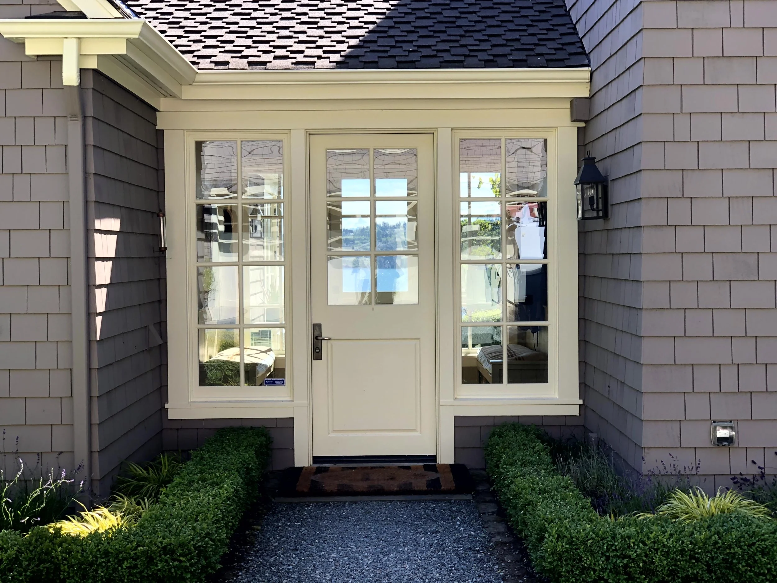 Front view of a house entrance with beige frame porch, large windows, a white door, surrounding purple shingles, and a pathway with green shrubbery.