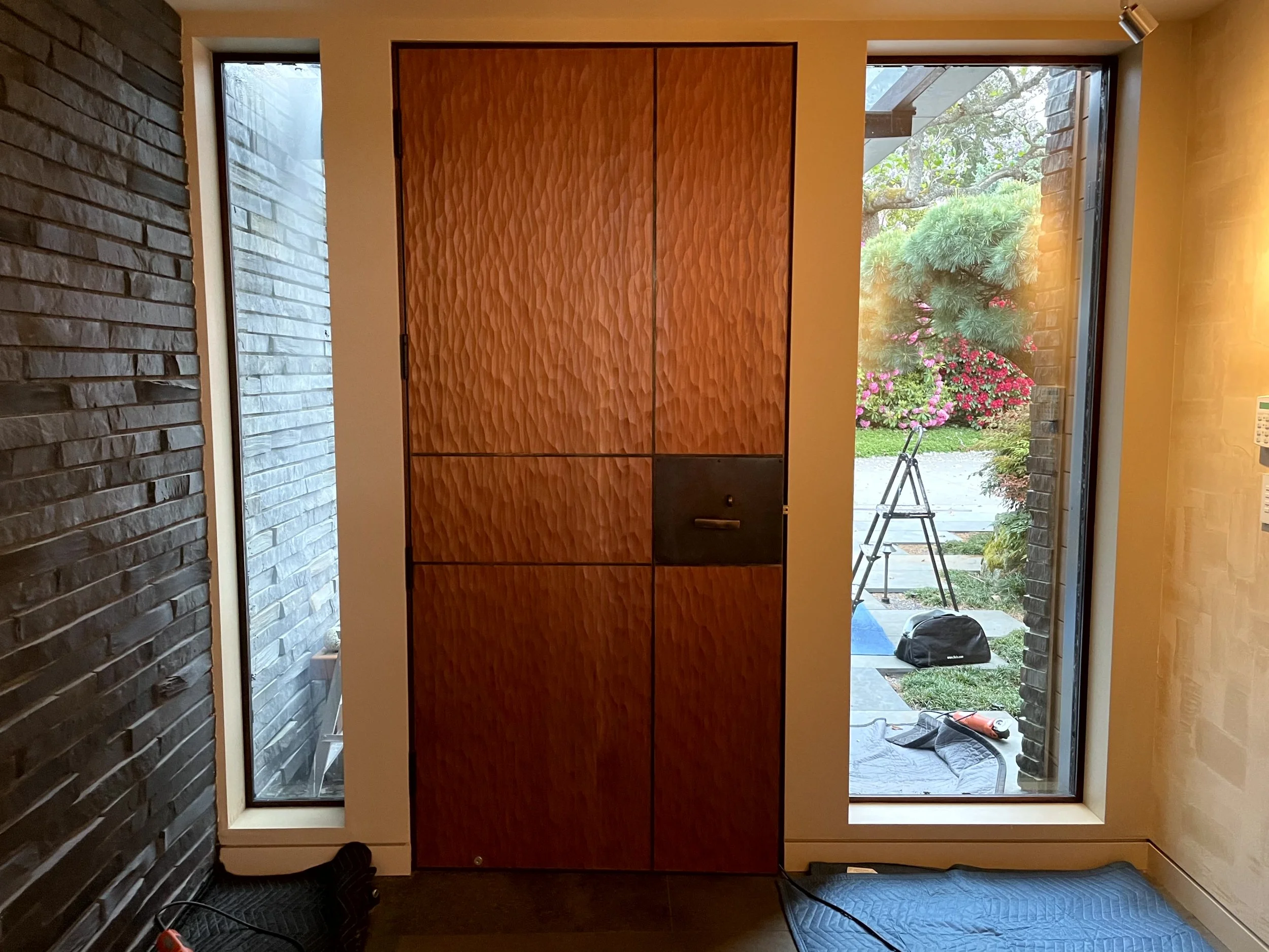 Interior view of a modern house showing a wooden cabinet or door in the center, flanked by narrow windows revealing a garden outside with flowering plants and patio tools.