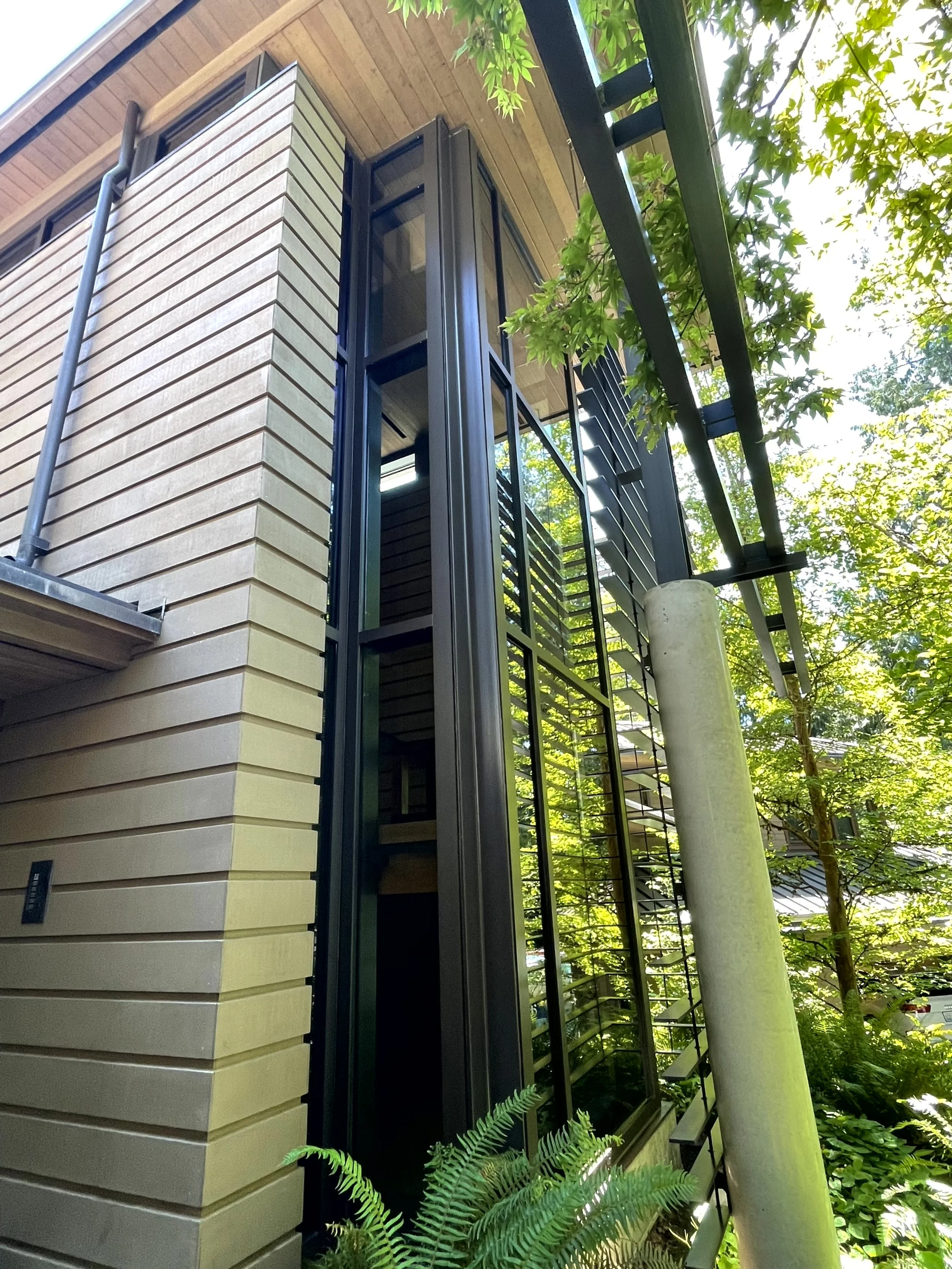Exterior view of a modern building with beige brick walls, black metal framing, and glass windows, surrounded by green trees and plants.