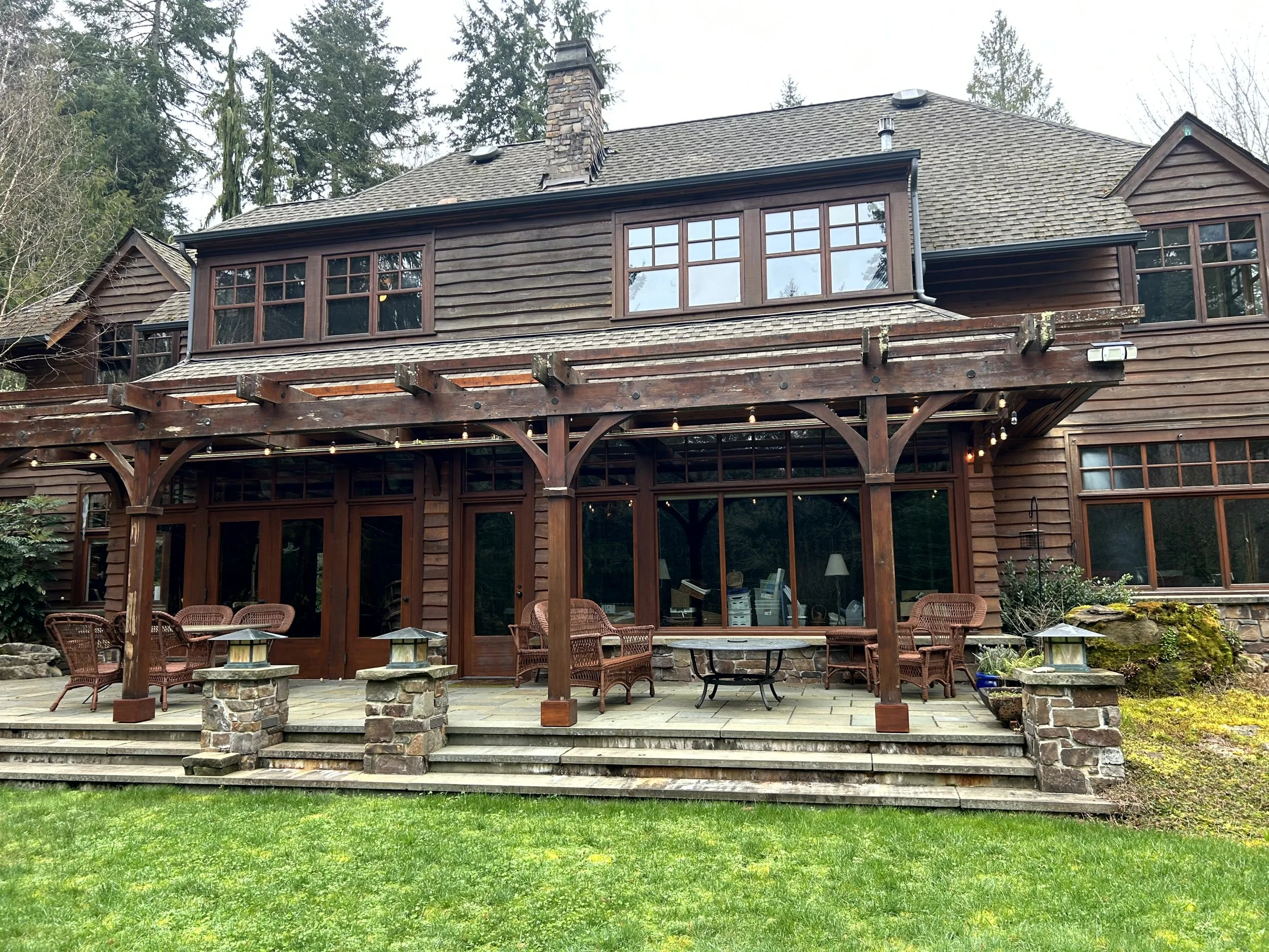 Rear view of a two-story wooden house with a large covered patio, outdoor seating, and string lights, surrounded by green grass and trees.
