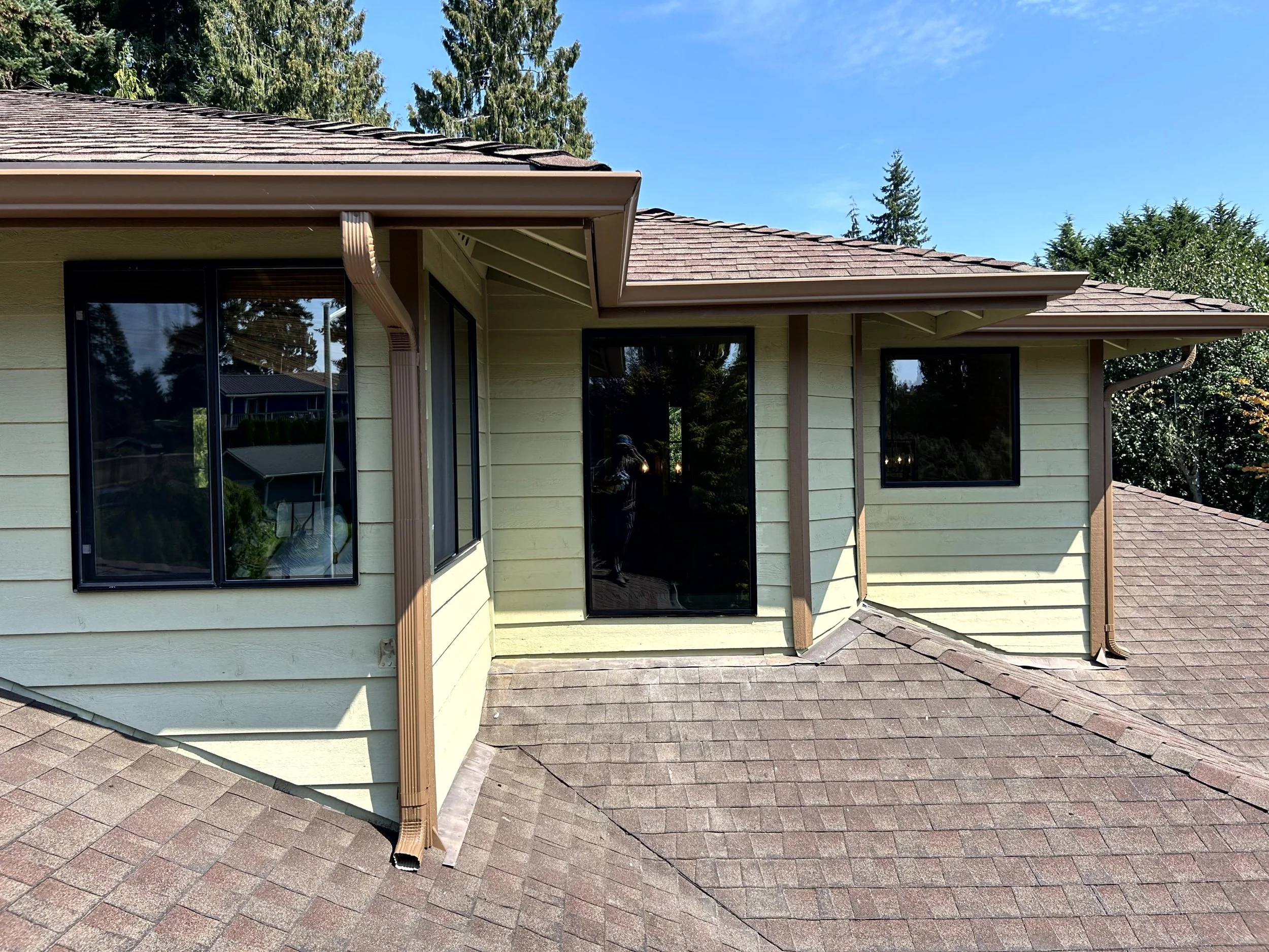 A beige house with brown roof shingles and black framed windows, surrounded by green trees and shrubs, under a blue sky.