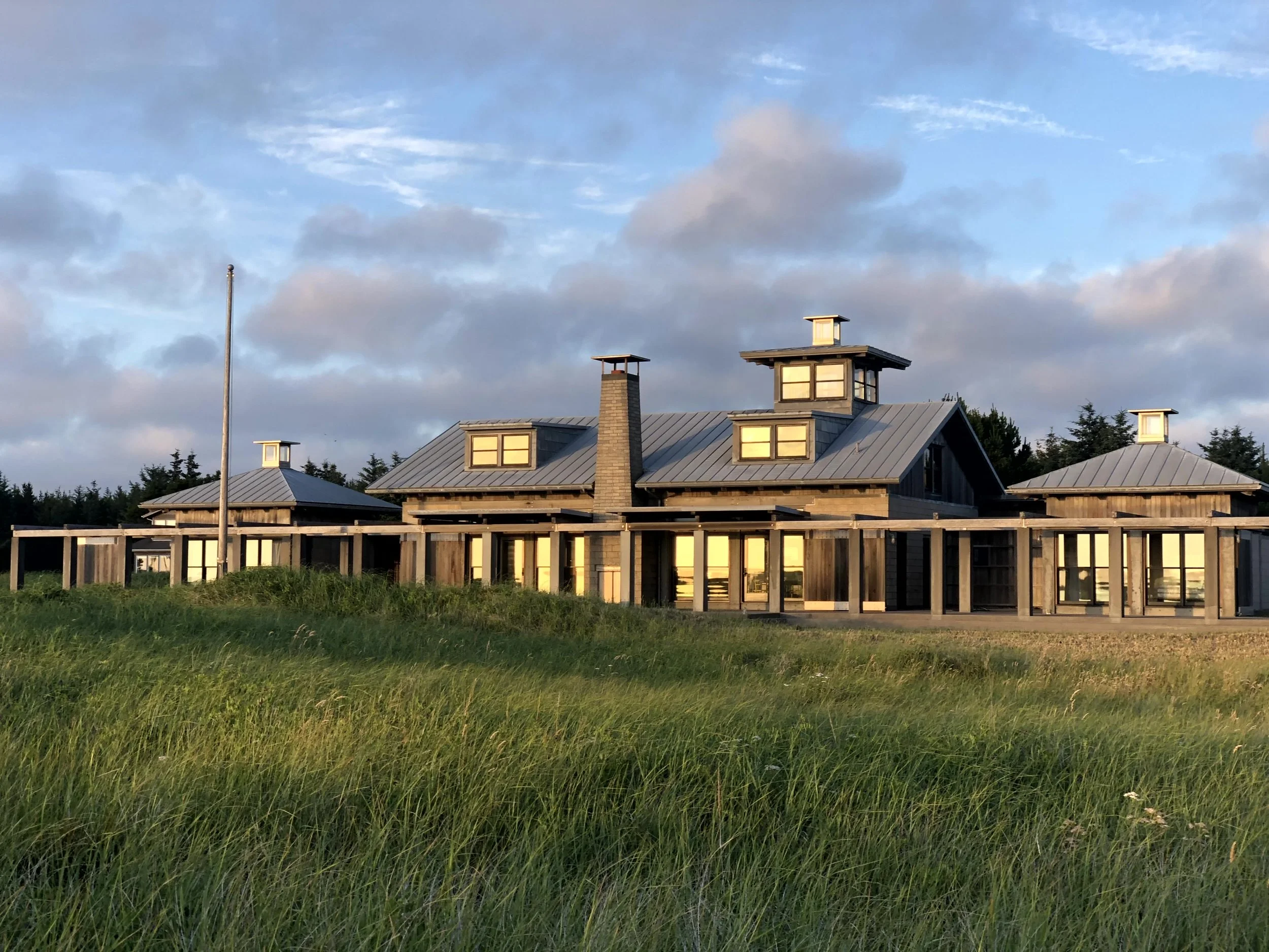 A rustic house with multiple pitched metal roofs and large windows, surrounded by a wooden fence, set in a grassy field during sunset with partly cloudy sky.