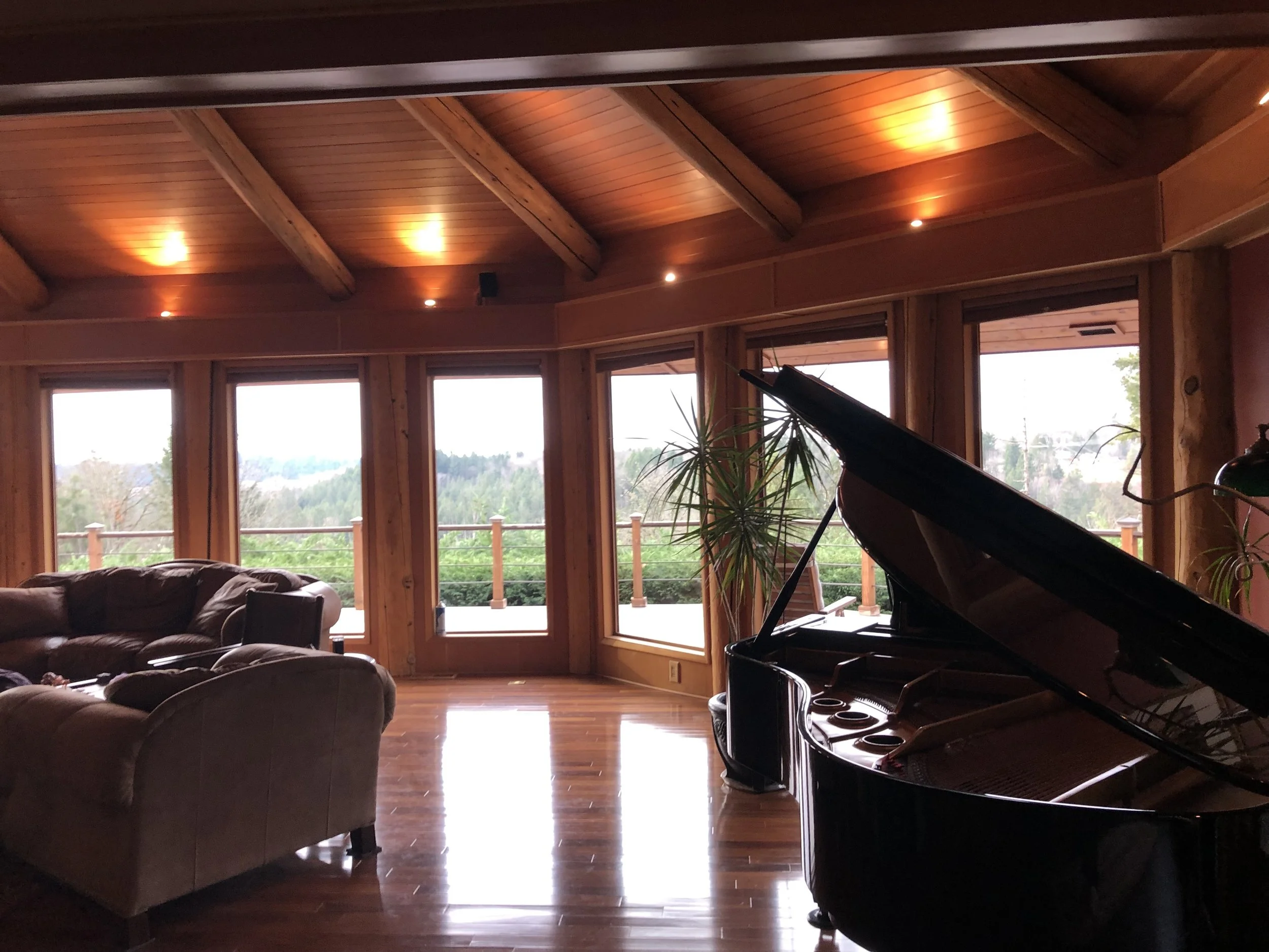 Interior of a living room with large windows overlooking a wooded landscape, wooden ceiling with exposed beams, a grand piano, and a seating area with leather sofas and plants.