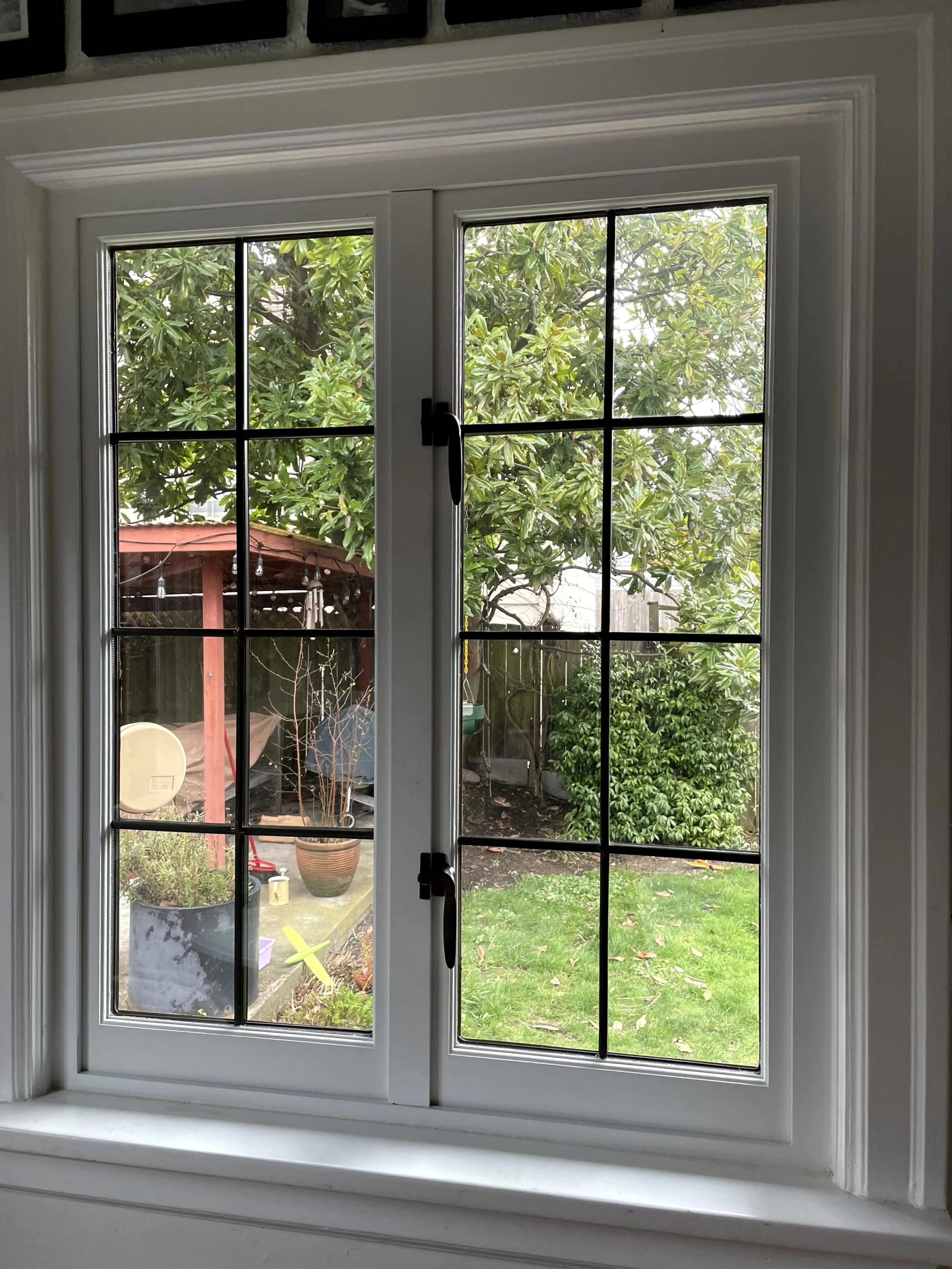 View through a two-pane white-framed window showing a backyard with trees, grass, a red shed, potted plants, and various garden items.