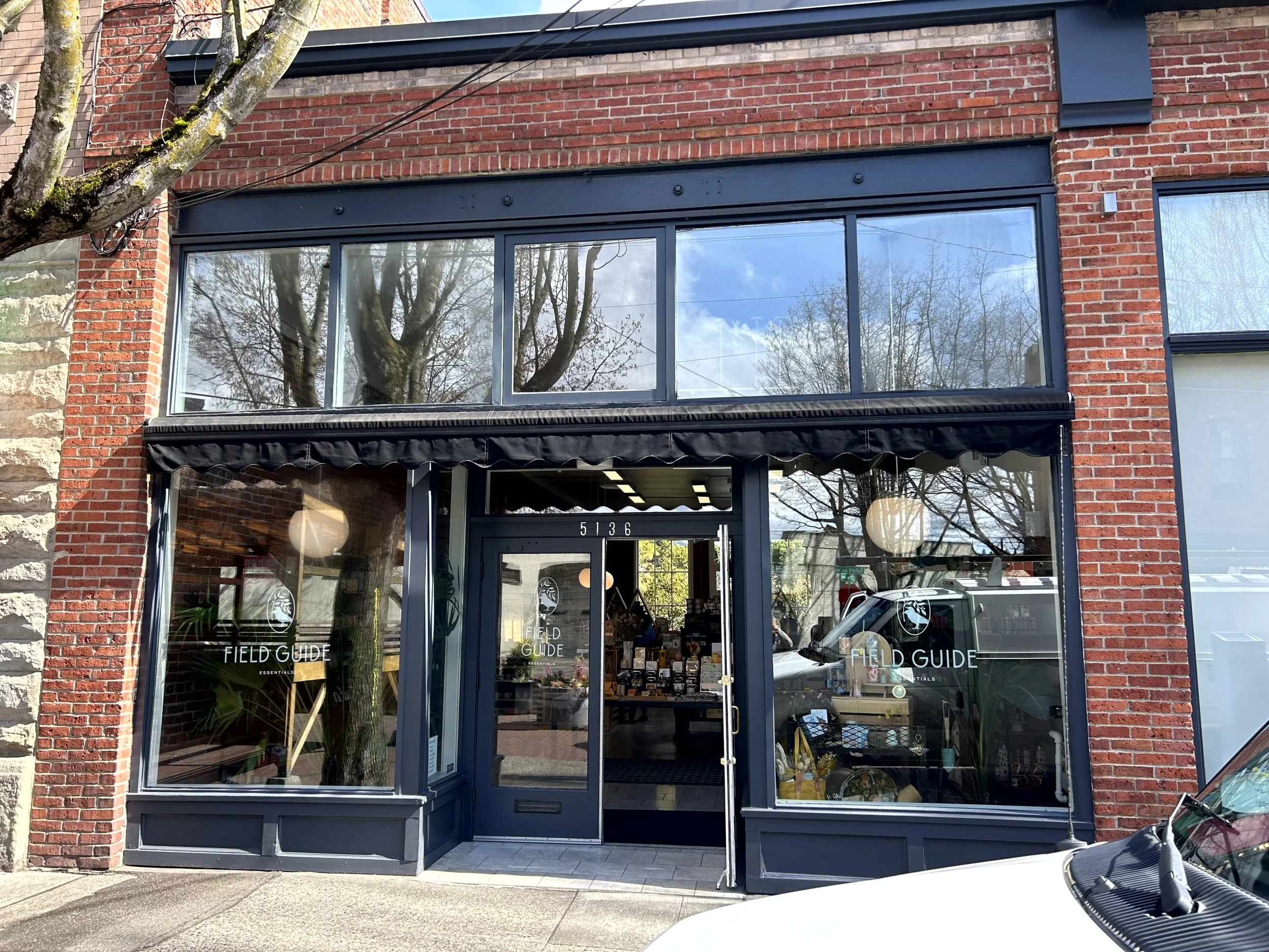 Storefront of a shop called 'Field Guide' with large glass windows and a door, brick exterior, and the store's logo on the windows. Reflection of trees and the sky can be seen in the windows.
