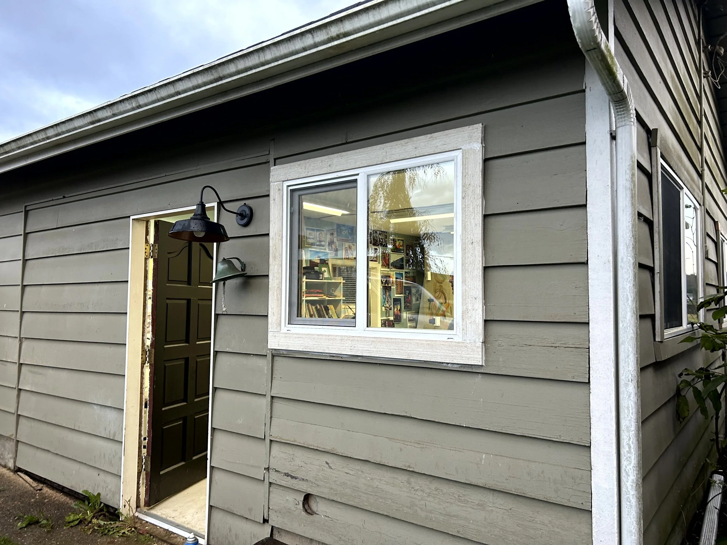 Exterior of a small building with gray horizontal siding, a window with a white frame, and an open door revealing bookshelves inside. There are two outdoor lights mounted next to the door.