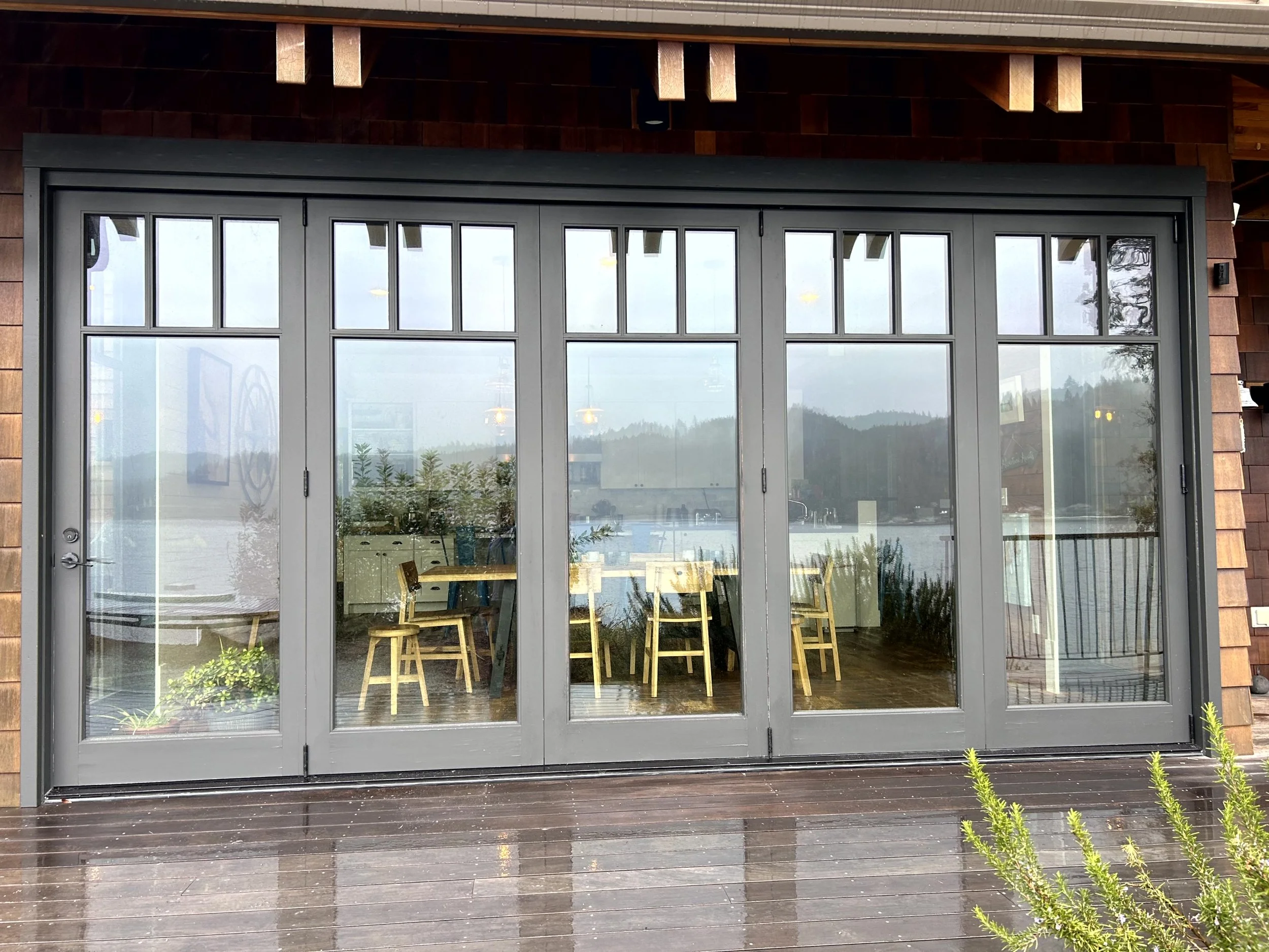 Large glass sliding door with a view of a lakeside landscape, wooden deck in the foreground, and a dining area inside with chairs.