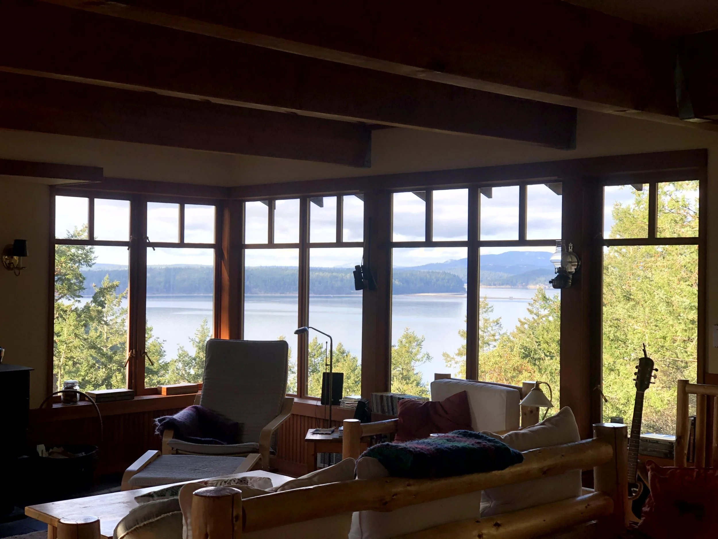 Interior view of a cozy living room with large windows showcasing a lake and forested landscape outside, with furniture including a sofa, chairs, and a guitar near the window.