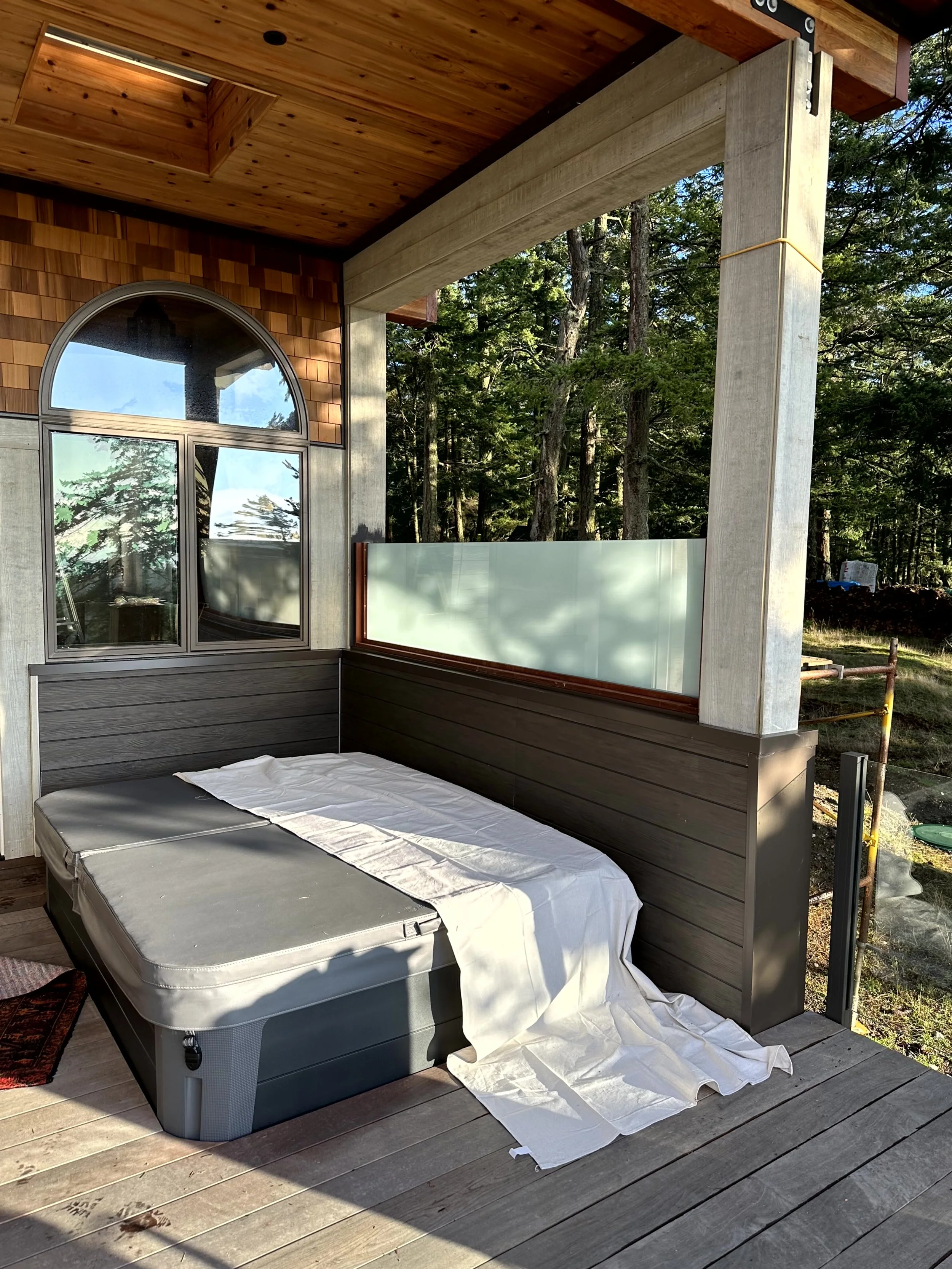 Outdoor balcony with a hot tub covered by a white cloth, surrounded by wooden flooring, a glass railing, and trees in the background.
