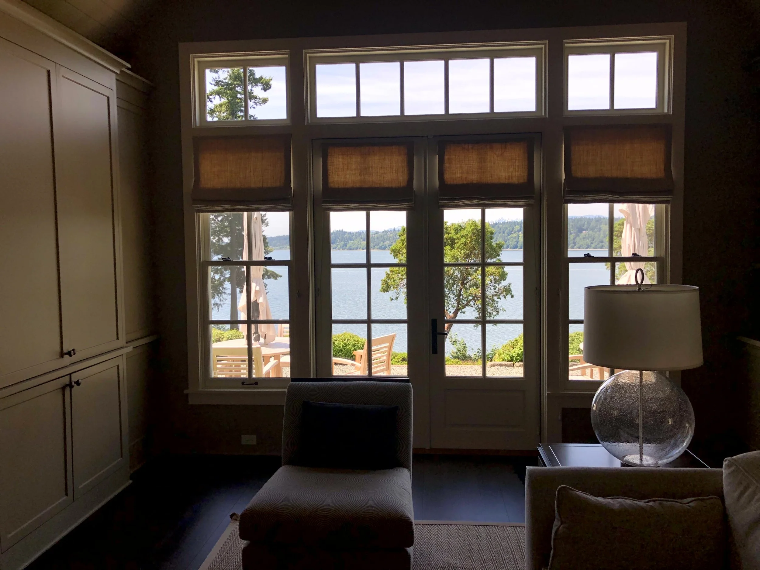 View of a lake through large glass doors and windows, with outdoor chairs and umbrellas on a patio, and greenery on the shore.