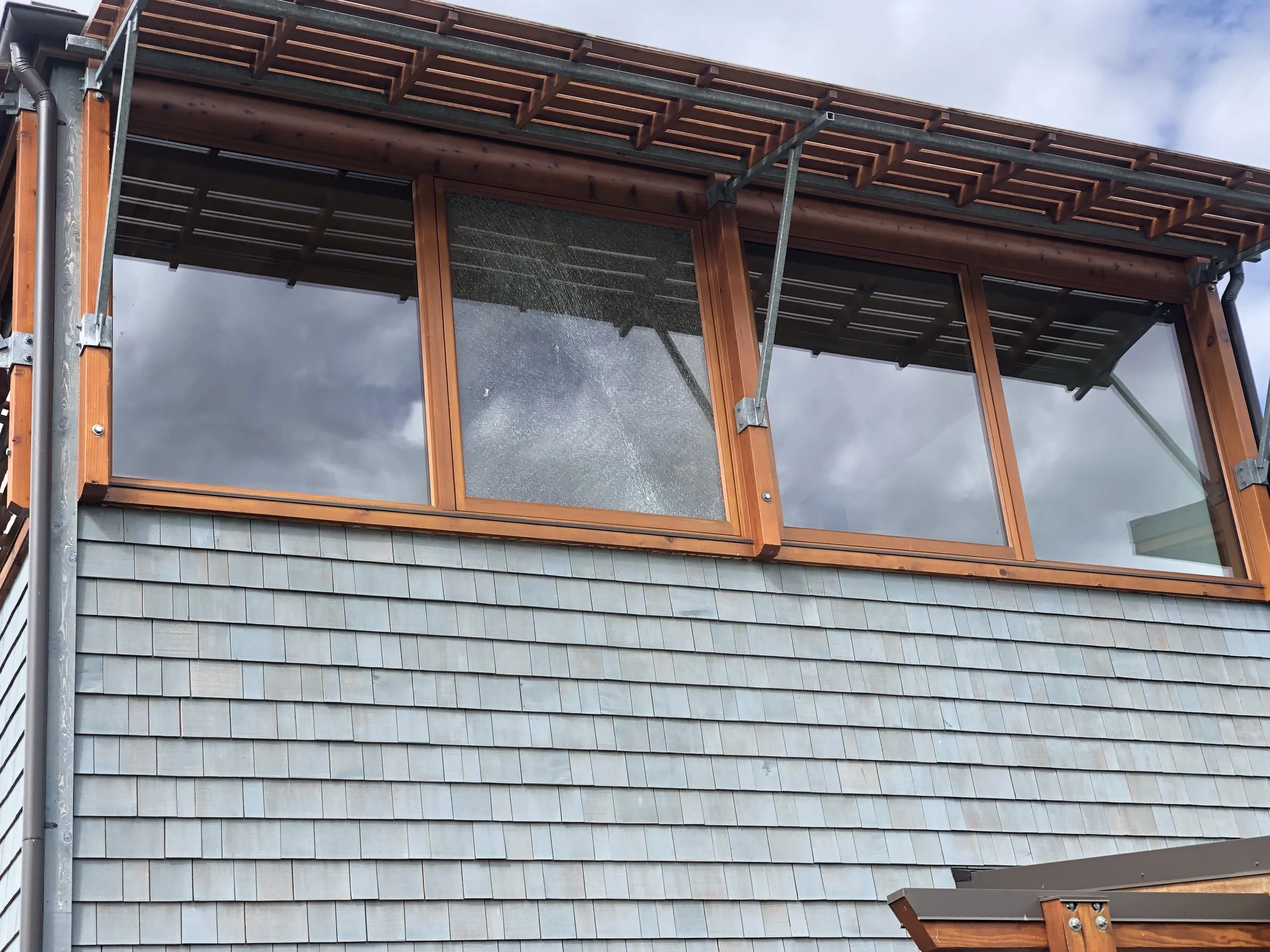 Close-up of a building's exterior showing large glass windows, a wooden awning, and gray shingle siding, with cloudy sky reflected in the windows.