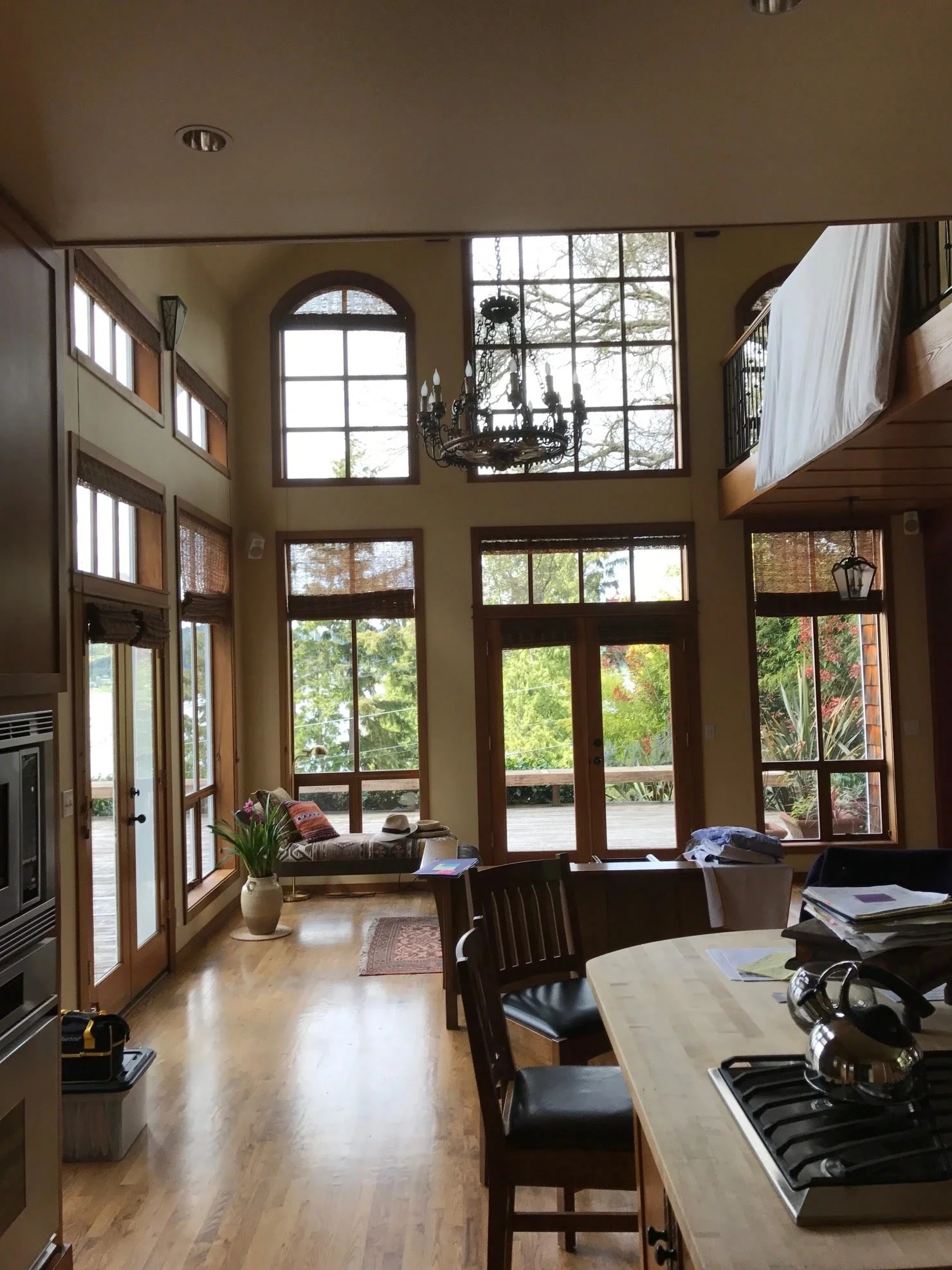 Interior of a sunlit living room with large windows, a chandelier, a bench with pillows, a potted plant, and a wooden dining table with chairs.