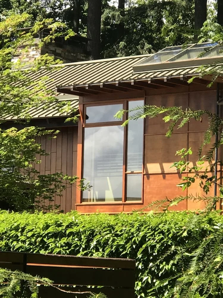 Close-up of a modern house with large glass windows, wooden exterior walls, and a green metal roof, surrounded by lush trees and shrubs.
