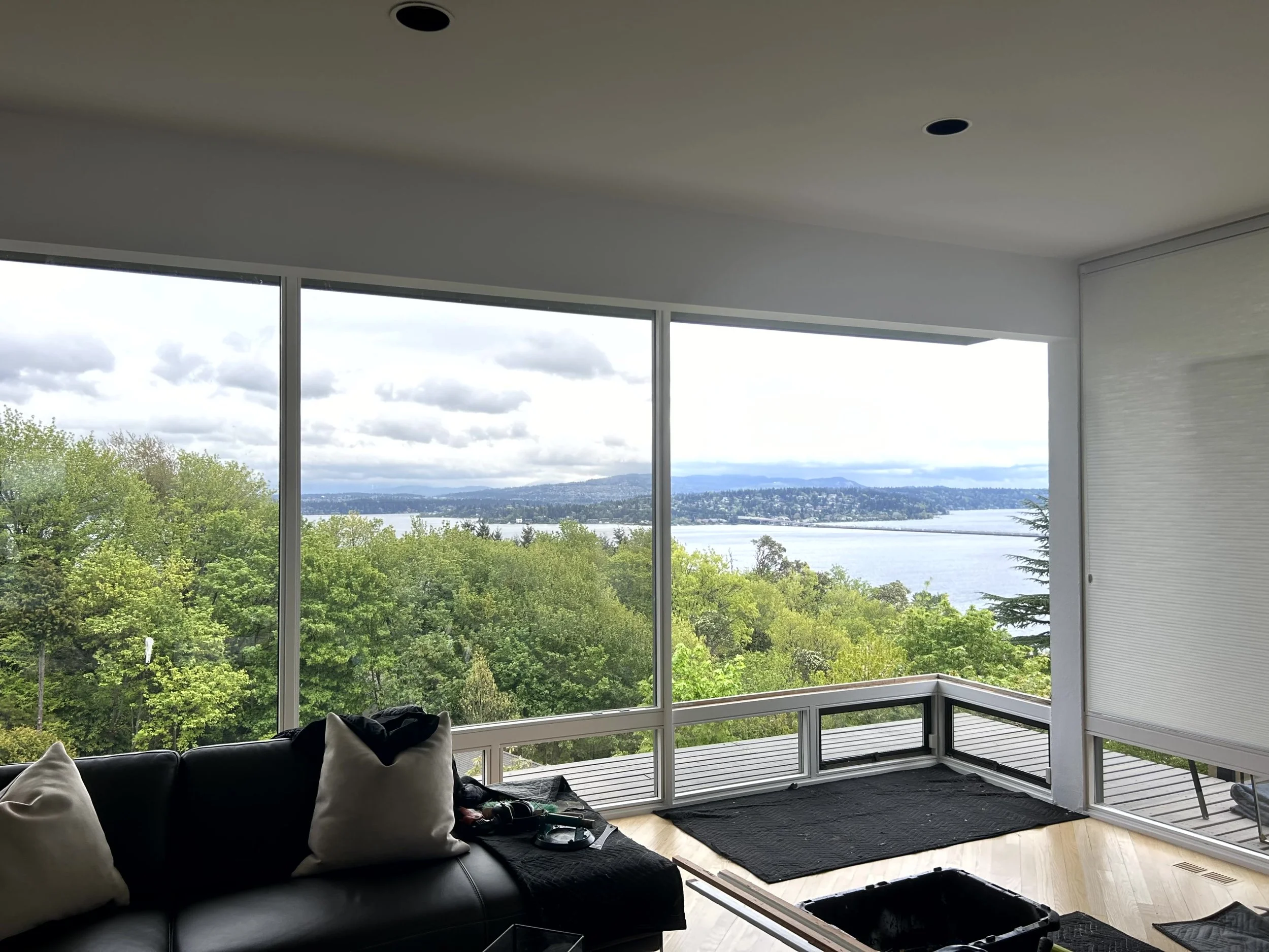 Living room with black leather sofa and large windows showcasing a view of trees, water, and distant hills under a cloudy sky.