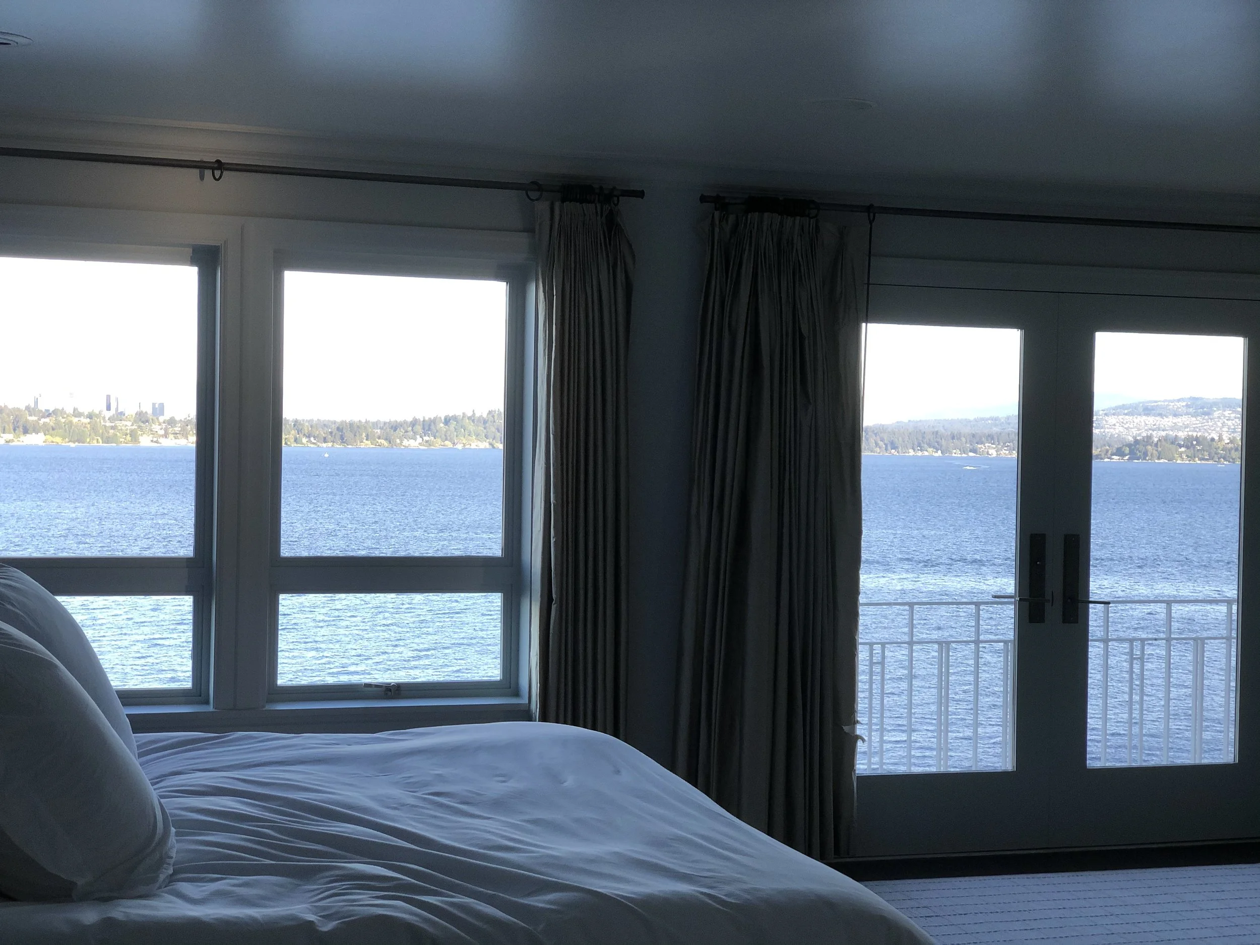 Interior view of a bedroom with a large bed and a window and glass door overlooking a body of water, with city buildings and mountains in the distance.