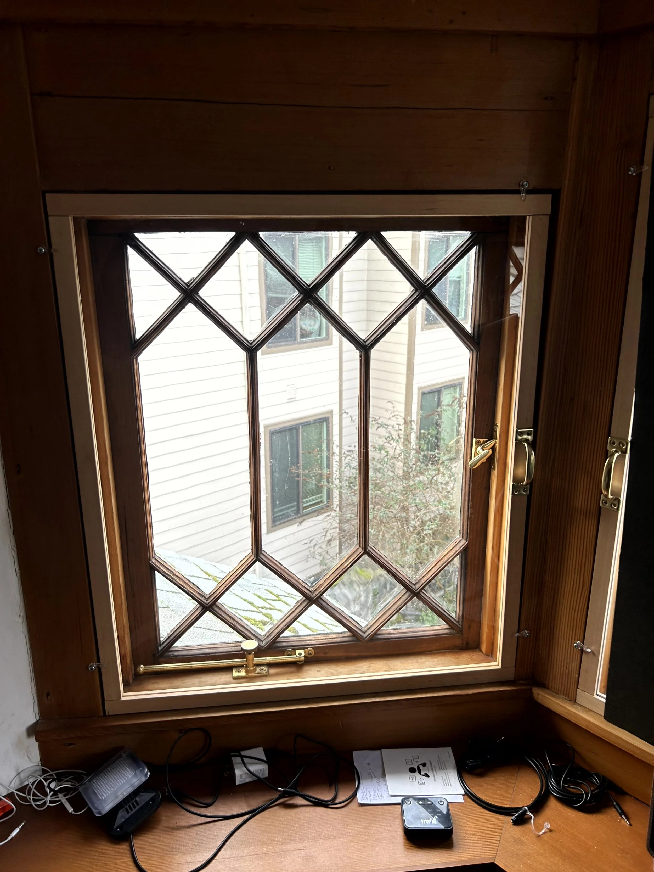 Wood window with diamond-shaped glass panes, partially open with a handle, showing a neighboring building with beige siding and windows, and some greenery outside.