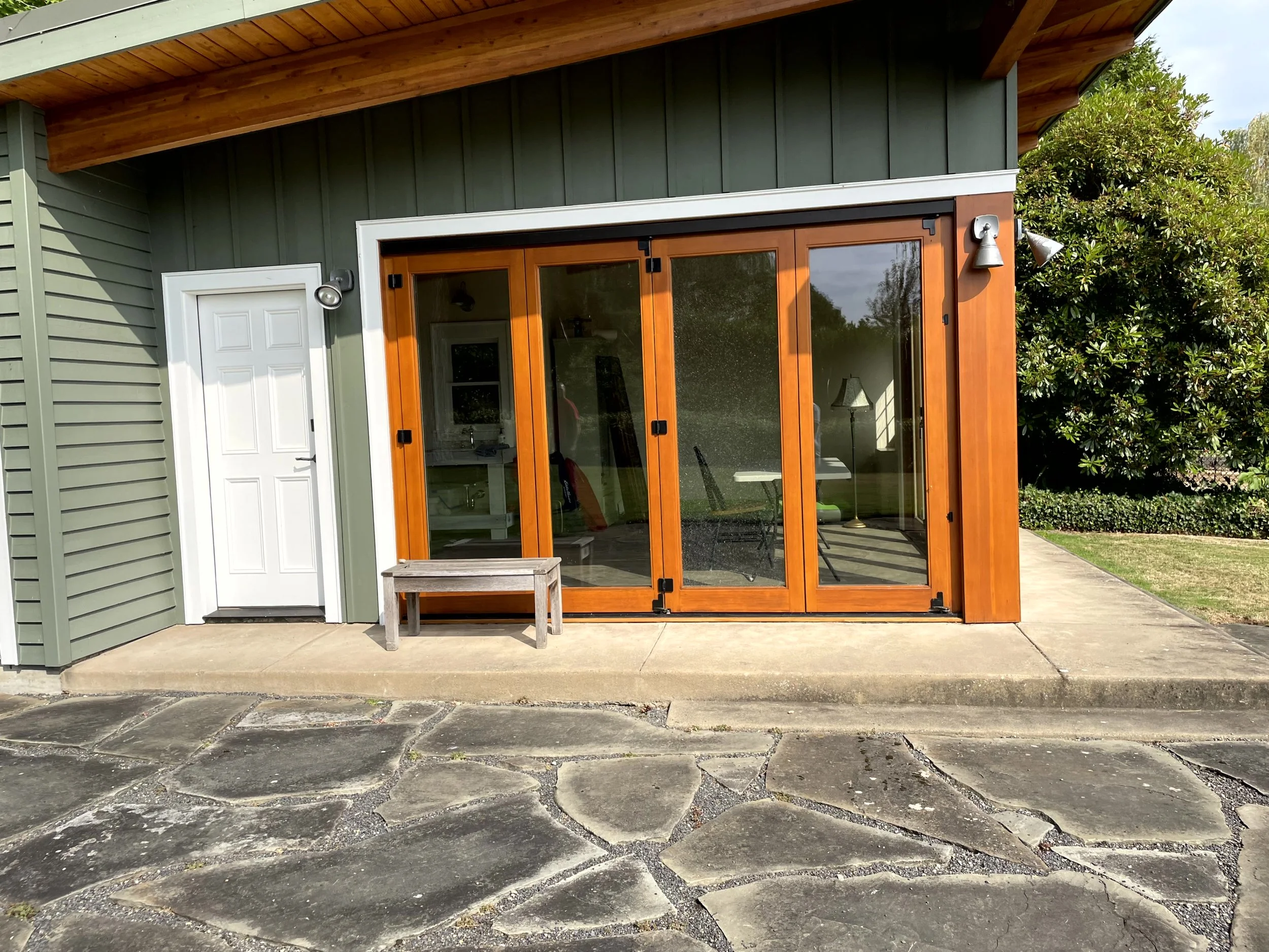 Exterior view of a house with a green siding wall, a white door, and large wooden sliding glass doors by a concrete porch and stone pavement. There are green trees and a lawn in the background.