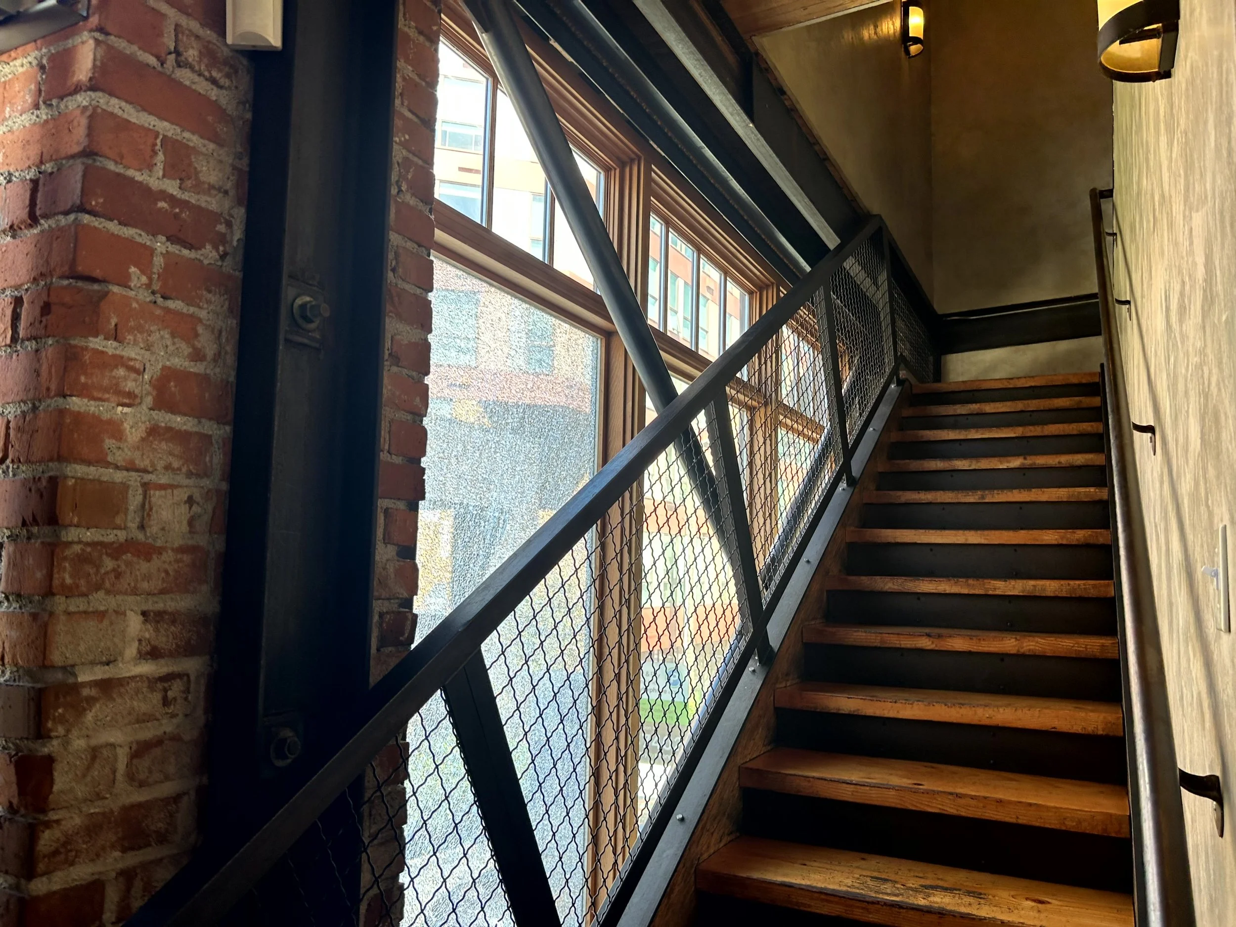 Interior staircase with wooden steps, metal railing, brick and yellow walls, large window letting in natural light.