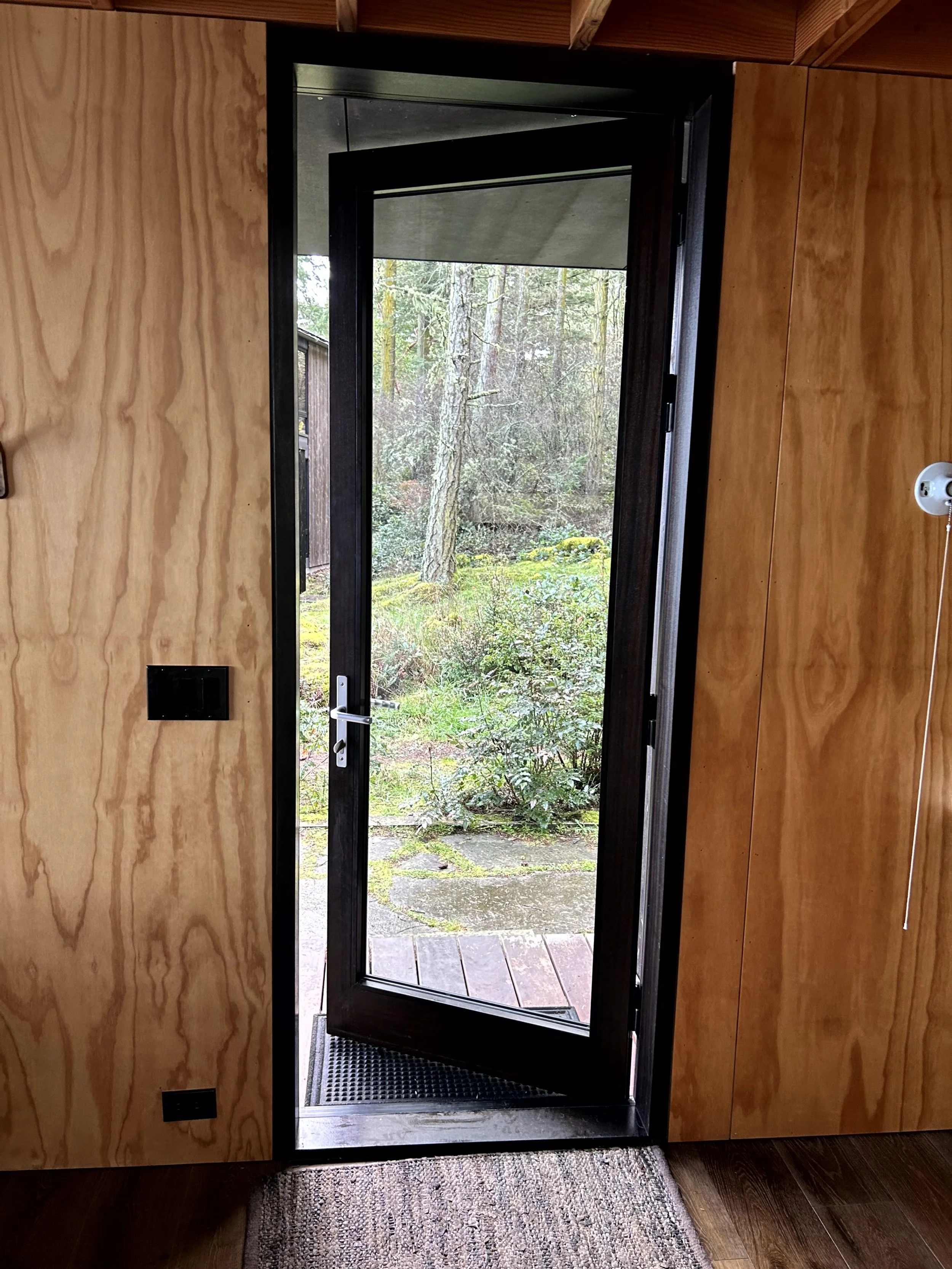 An open black-framed glass door leading to a muddy garden path with trees and bushes outside.