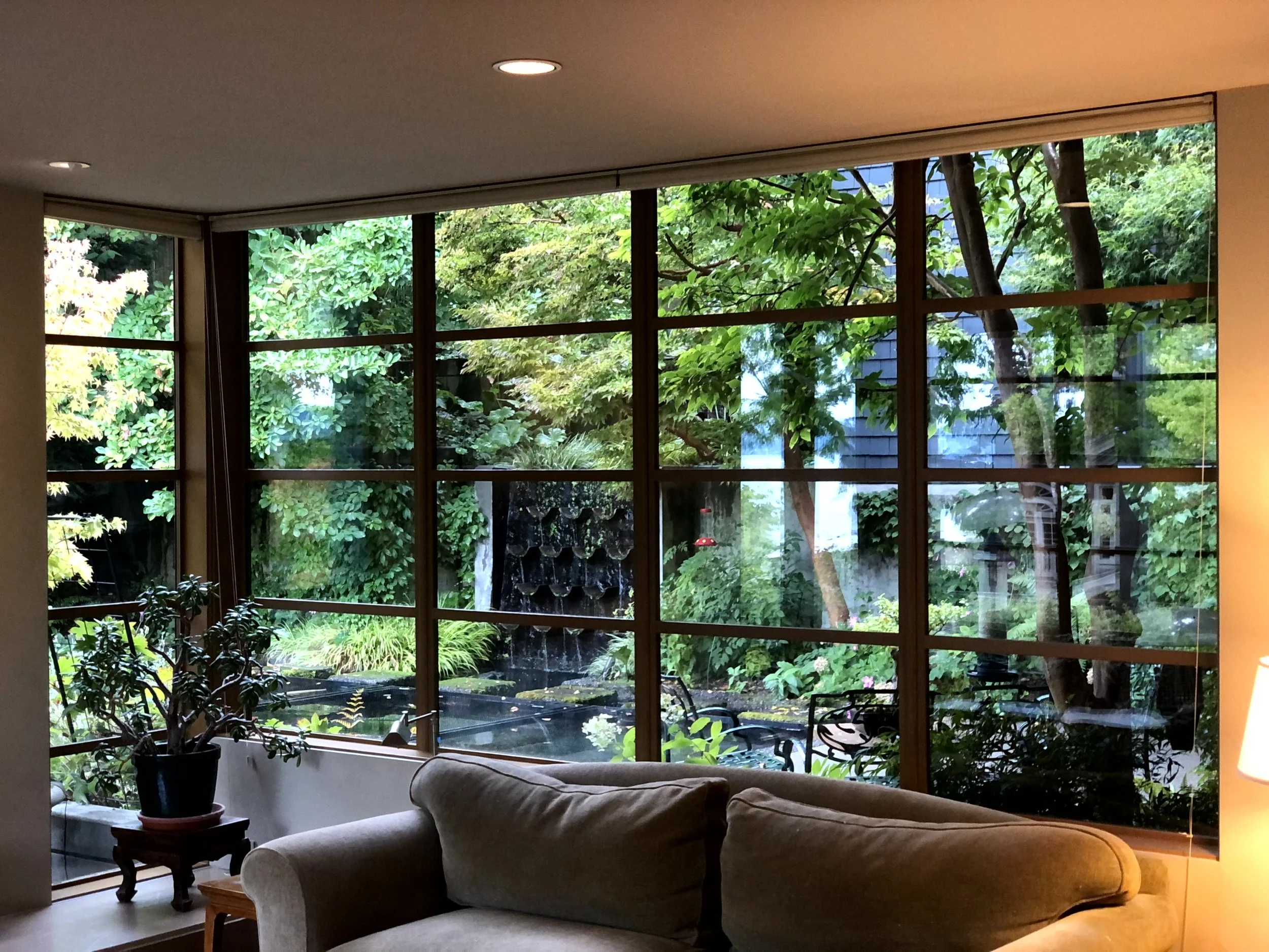 Living room with large window looking out onto a lush green garden with trees, plants, and a small pond, a beige sofa, a potted plant, and a lamp.