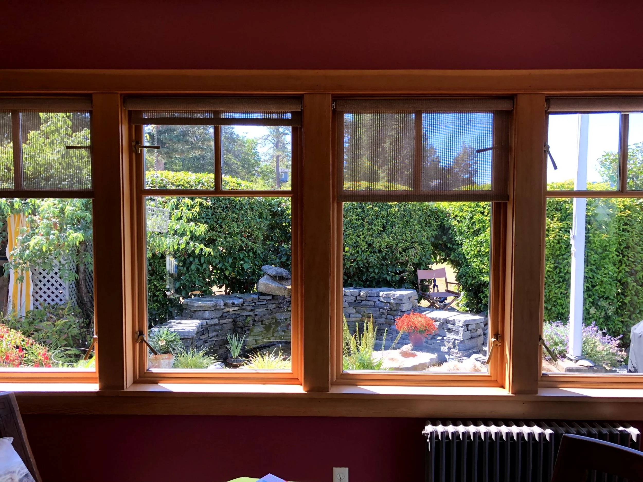 View of a garden through three wooden framed windows with screens, showing plants, a stone wall, and a bench outside.