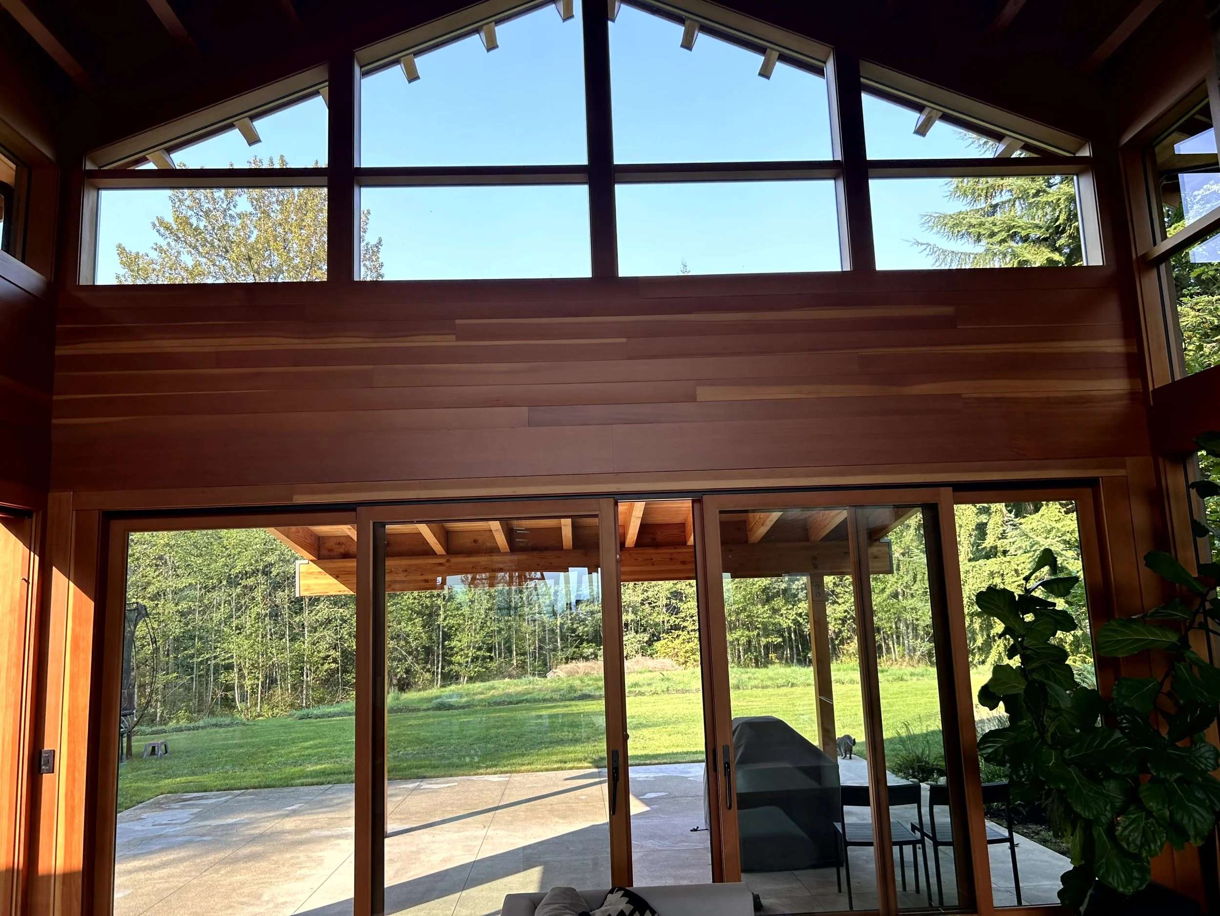 Interior view of a modern home with large glass sliding doors leading to a backyard with green grass and trees, and floor-to-ceiling windows allowing natural light inside.
