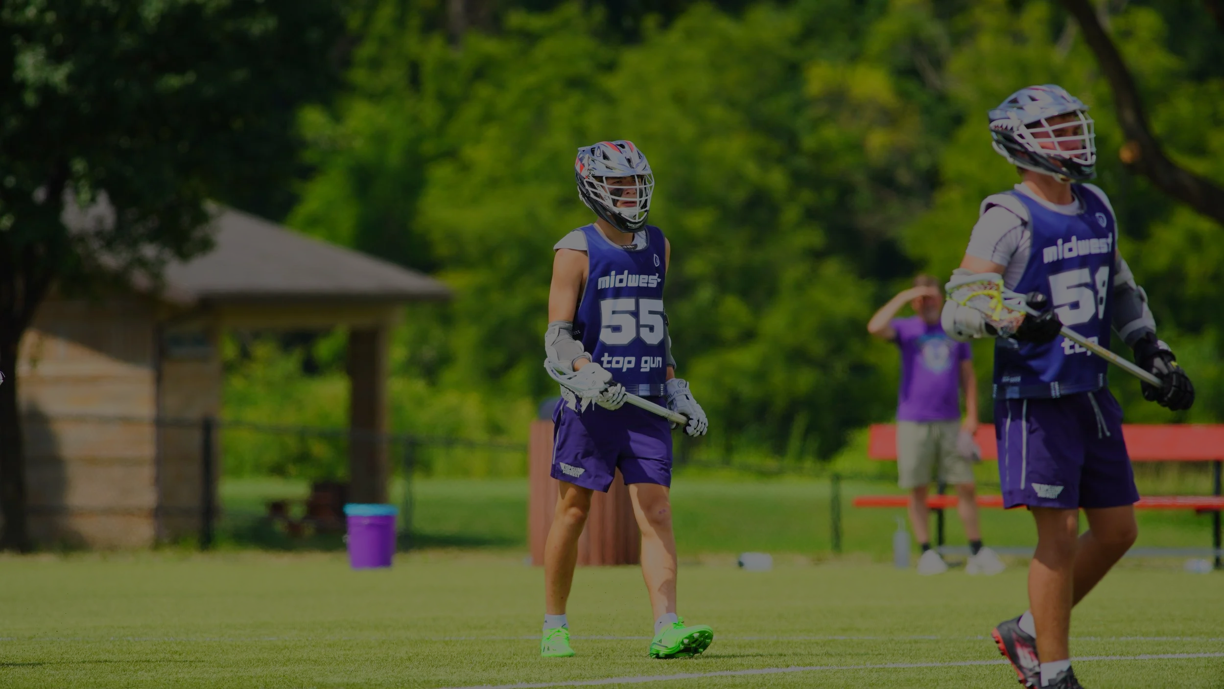 Two youth lacrosse players wearing helmets and purple jerseys with the word "midwest" on front, playing on a green field, with a person in a purple shirt in the background.