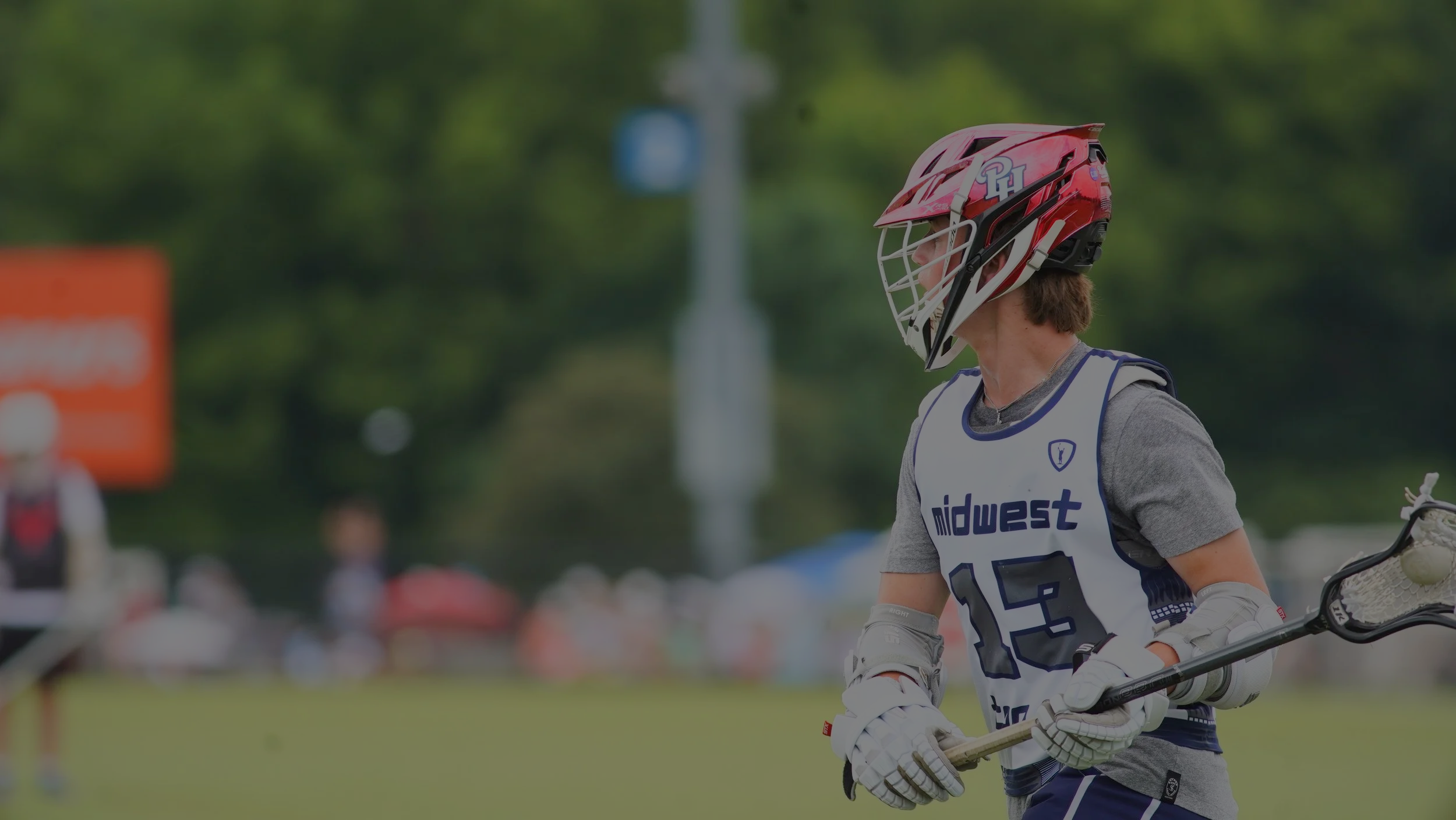 A young lacrosse player wearing a red helmet, a gray T-shirt, white gloves, and a white jersey with the number 13 participates in a game on a grassy field. The background is blurred with other players, tents, and trees.
