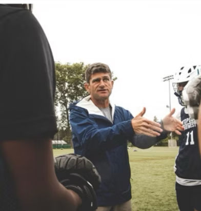 A man in a blue jacket speaking and gesturing with his hands during a conversation on a sports field with football players.