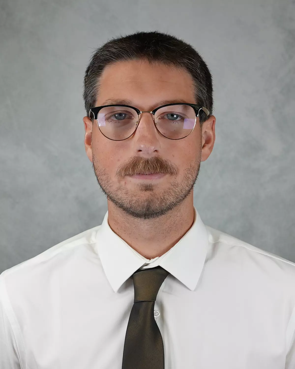 A man with short dark hair, glasses, and a beard and mustache wearing a white shirt and a dark tie, standing against a plain gray background.