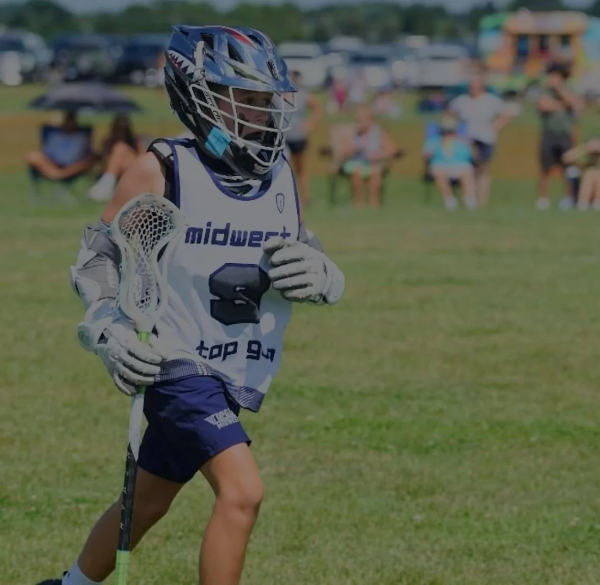 Lacrosse player running on the field wearing a helmet, gloves, and a jersey with 'midwest' and 'top 94' printed on it, holding a lacrosse stick. Spectators watch in the background.