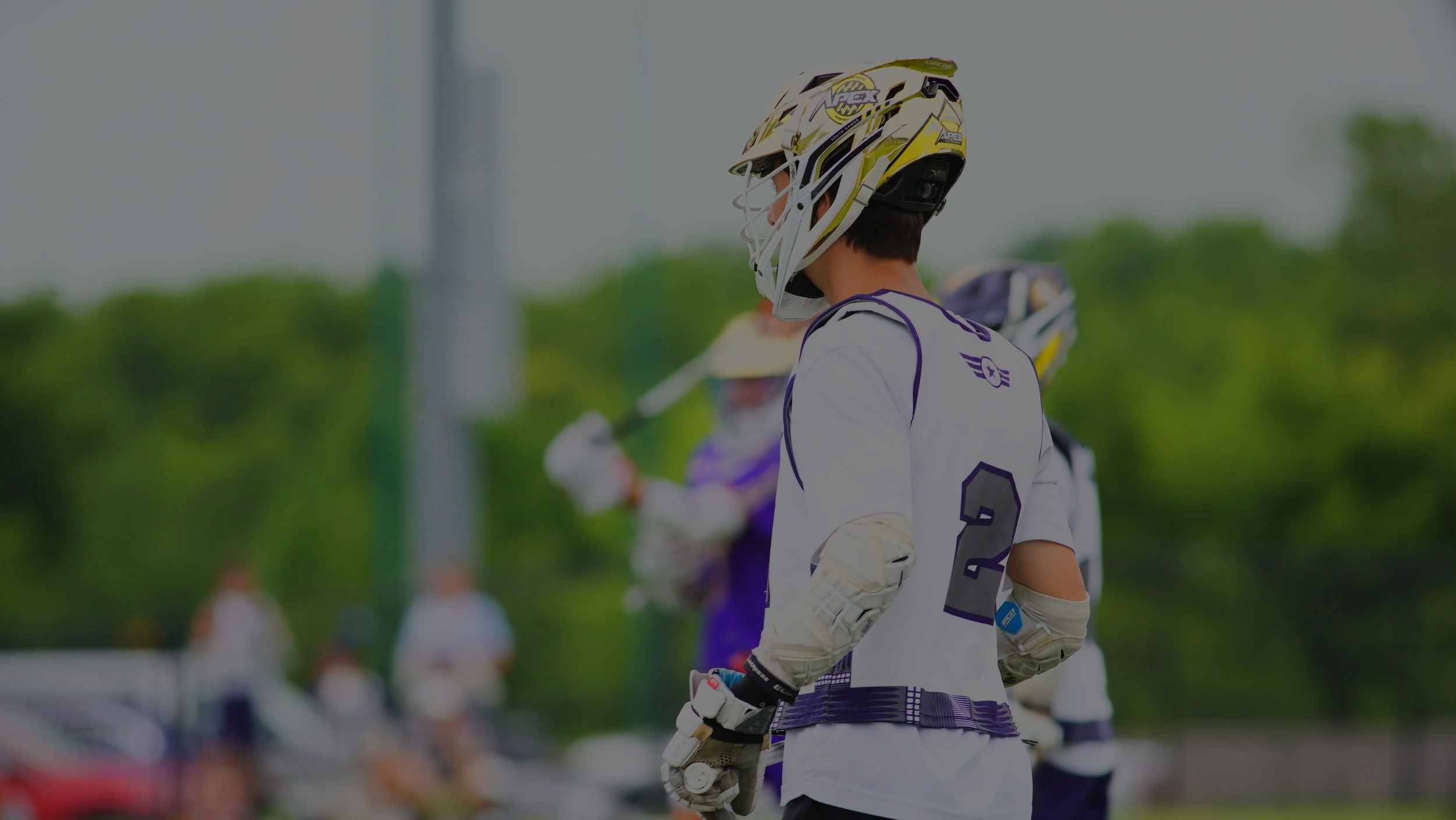 Lacrosse player in a white jersey and helmet with a yellow and black design, holding a lacrosse stick, standing on a field with green trees in the background.