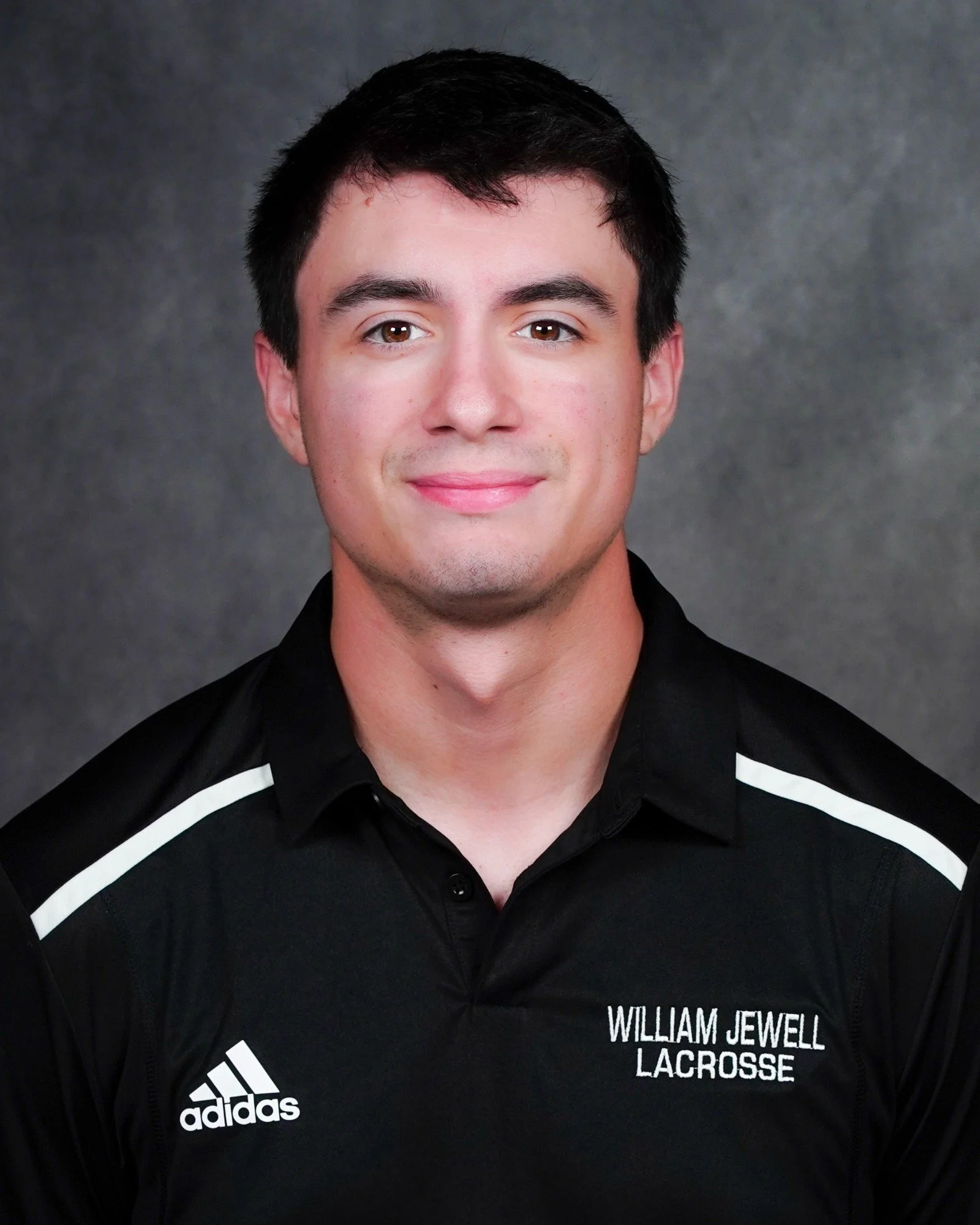 A young man with short dark hair and light skin, wearing a black Adidas sports shirt with white accents, featuring the text 'William Jewell Lacrosse' on the chest, smiling against a gray background.