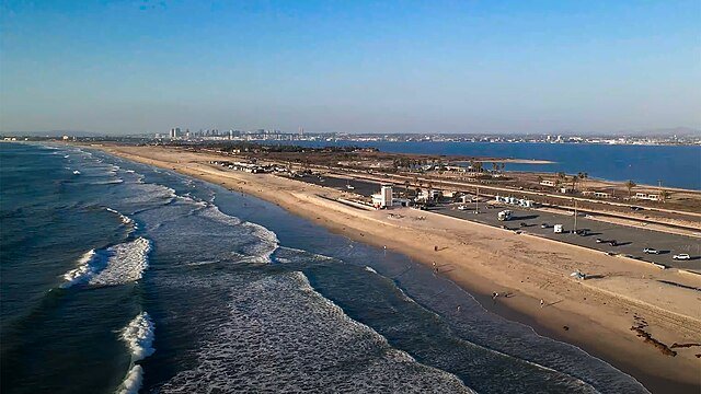 Beach with waves and a road parallel to the shoreline, with a cityscape in the background under a clear blue sky.