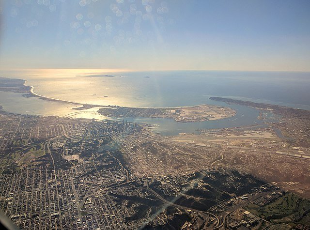Aerial view of a coastal city with a large harbor, residential neighborhoods, and the ocean in the background.