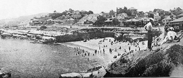 Historical black and white photo of a coastal area with a beach filled with people, and houses on the hillside in the background.