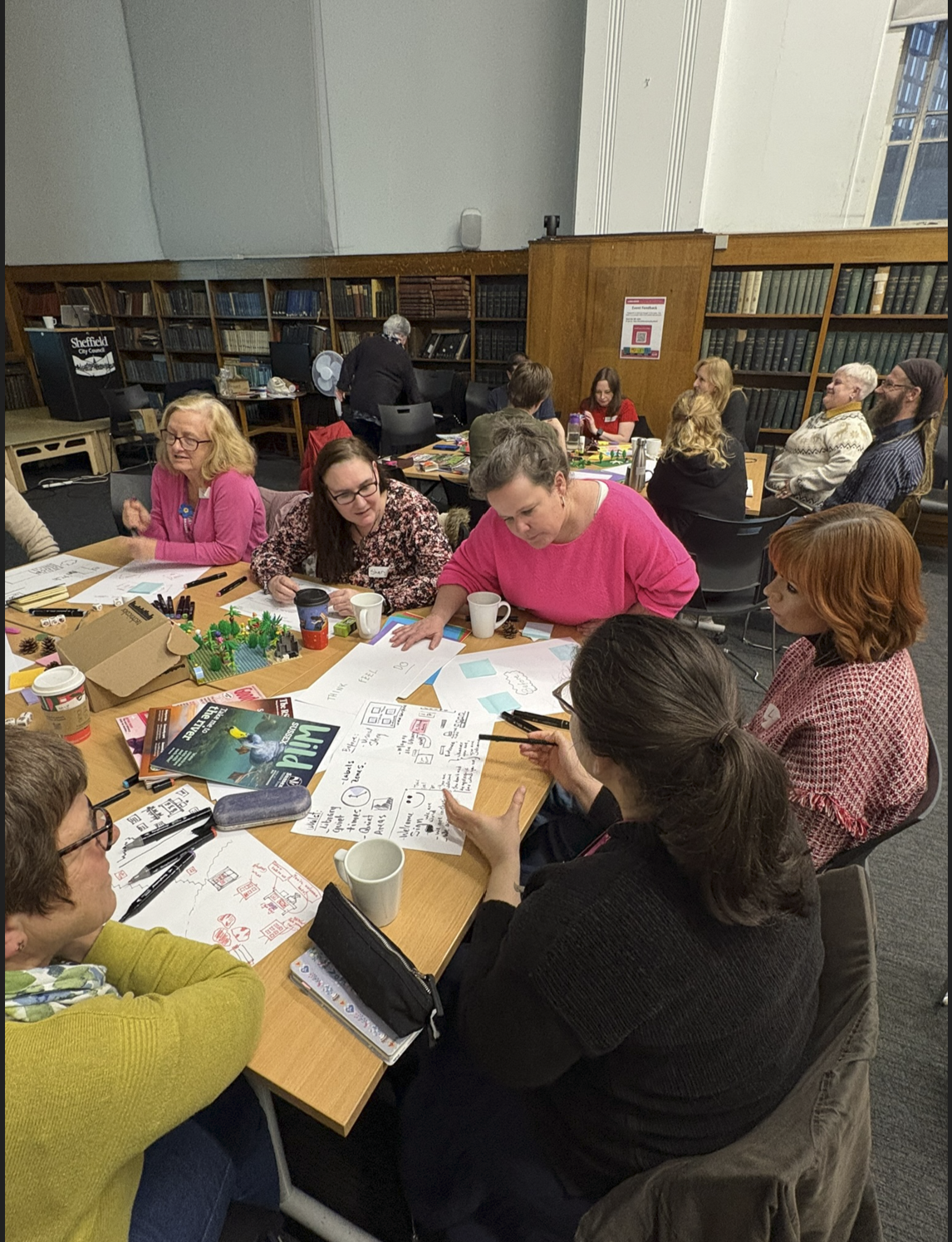 Sascha leading a creative workshop session with a group of people in a library setting, with worksheets and diagrams spread across a table.