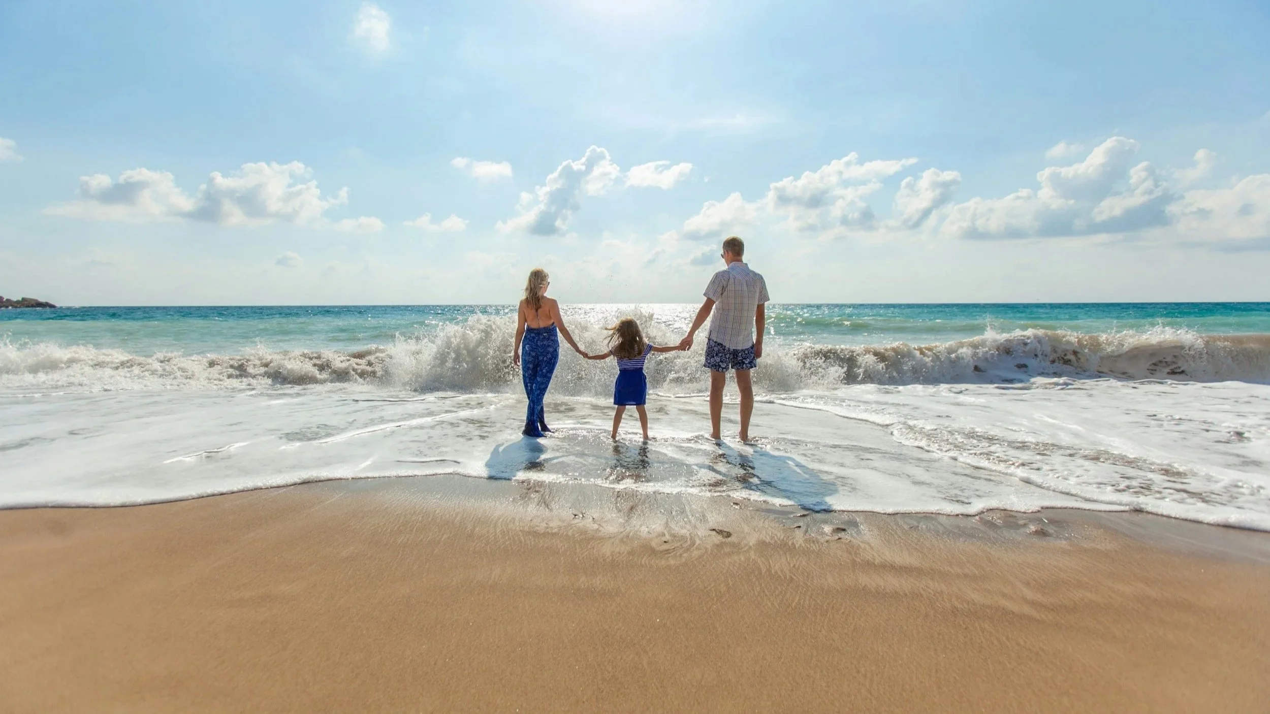 Family of four holding hands and walking into the ocean waves on a sunny beach.