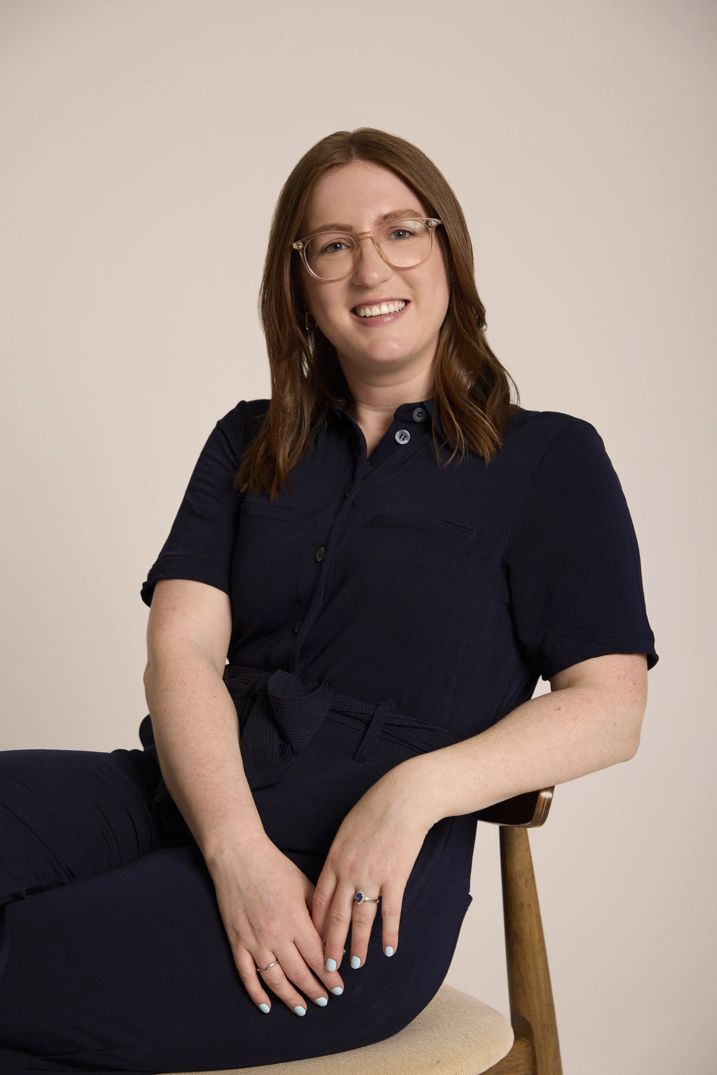 A woman with shoulder-length brown hair and glasses sitting on a beige chair, smiling at the camera, wearing a navy blue dress with rolled-up sleeves and a belt.