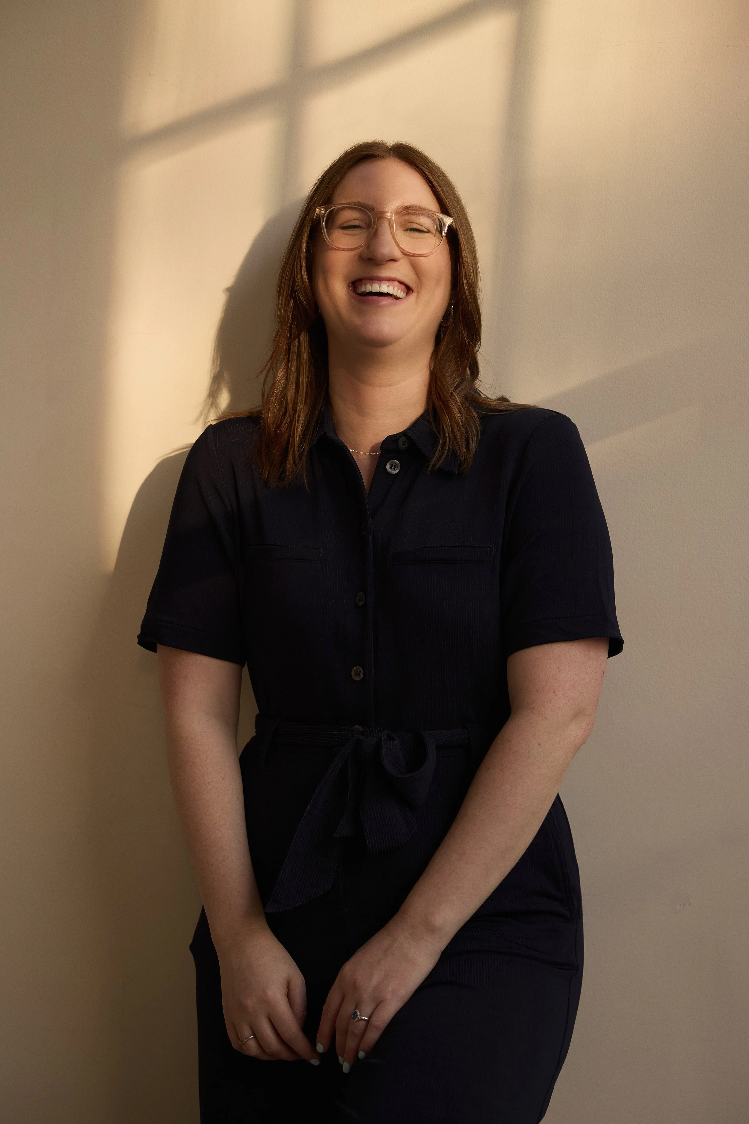 Woman with glasses smiling and laughing, wearing a navy jumpsuit, standing against a wall with shadows of window panes.