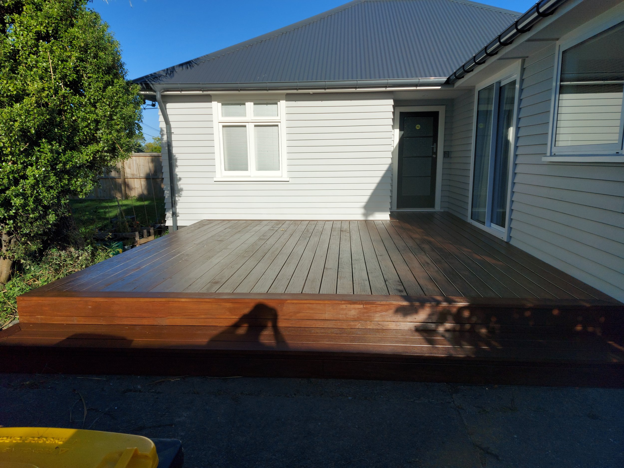 A newly stained wooden deck in front of a modern, light-colored house with a metal roof. The deck is clean and extends from the house's sliding glass door. The yard is partially visible with a tree on the left and a blue sky overhead in Ch