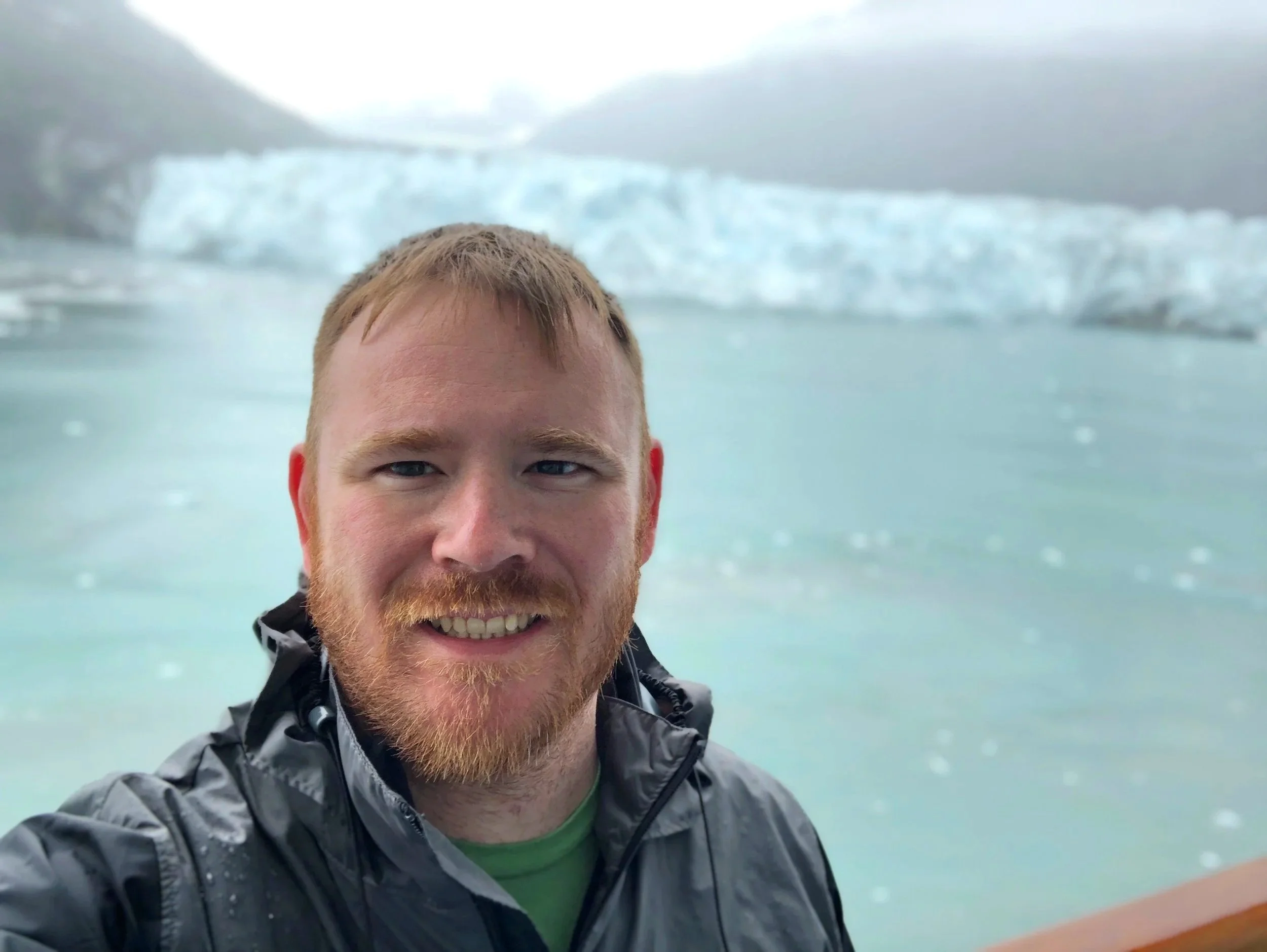 A man with a beard and short red hair taking a selfie in front of icy glaciers and water.  This is the founder of Modern Mens Clinic